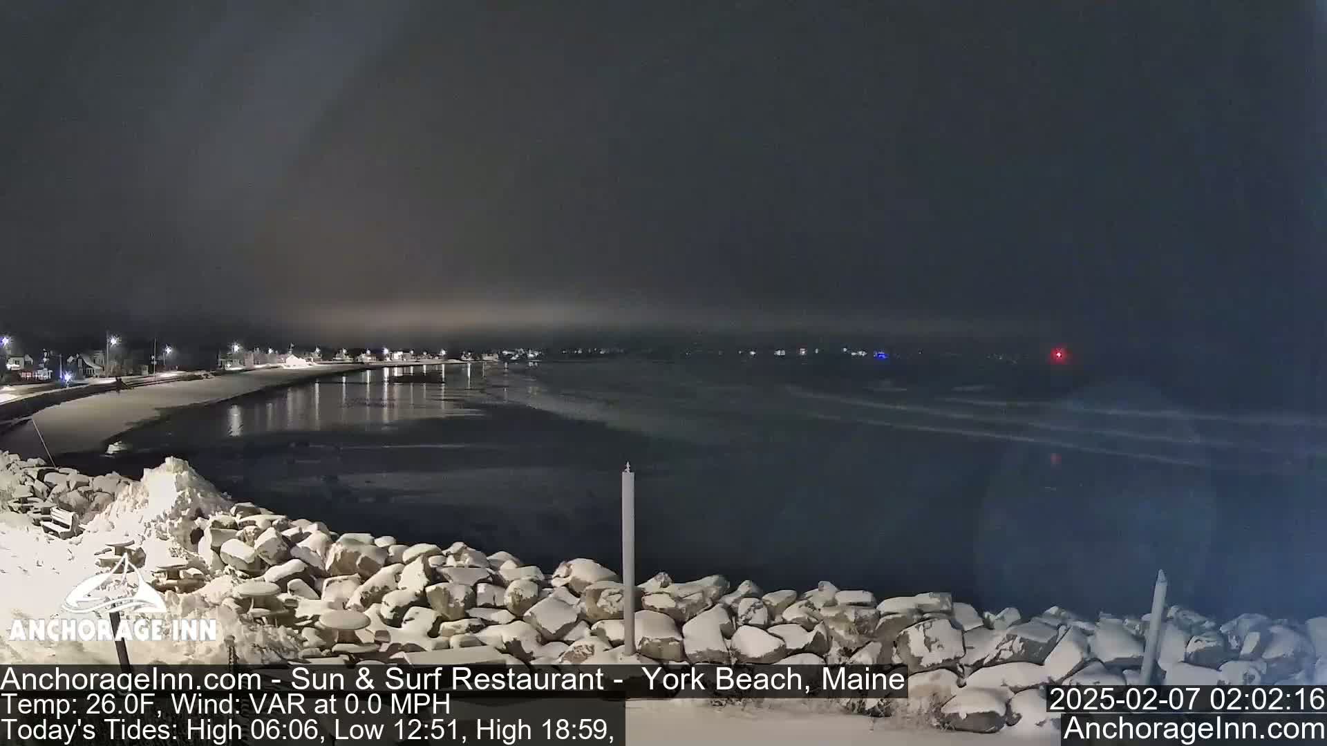 A snow-covered rocky shoreline at night overlooks a calm, dark body of water with a town faintly visible in the distance under a dark sky.