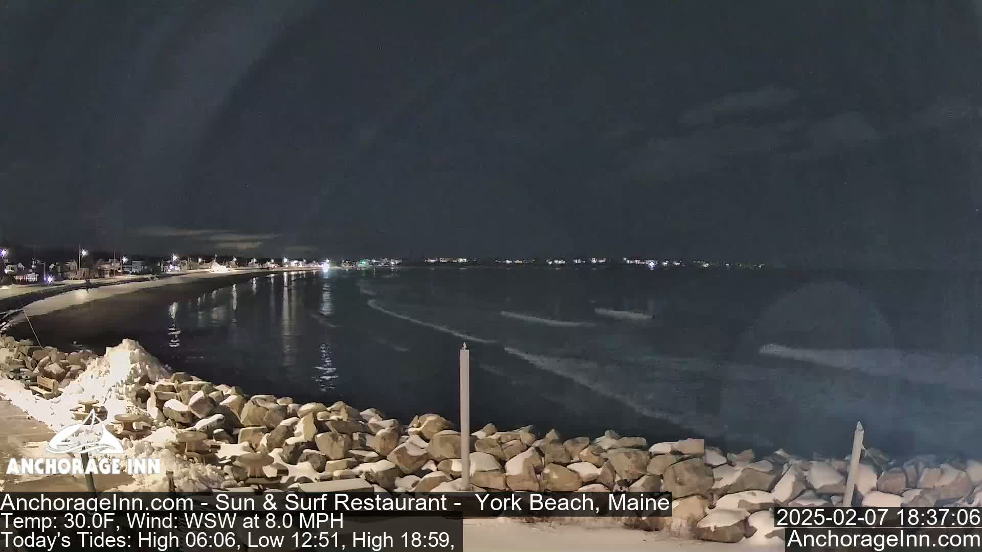 A nighttime view of a snow-dusted rocky shoreline and calm ocean under a dark sky.