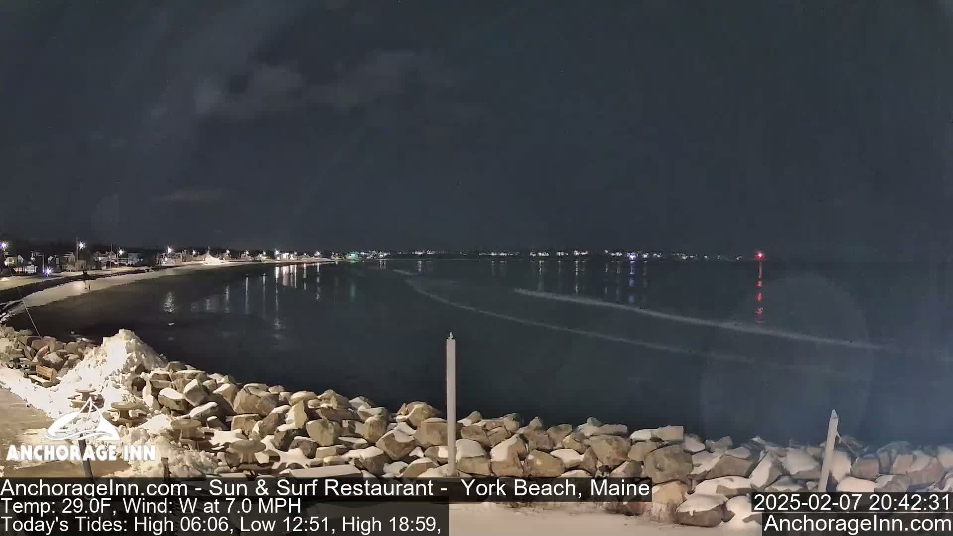 A nighttime view of a snow-dusted rocky shoreline and calm, dark water reflecting a few distant lights, under a mostly clear sky.