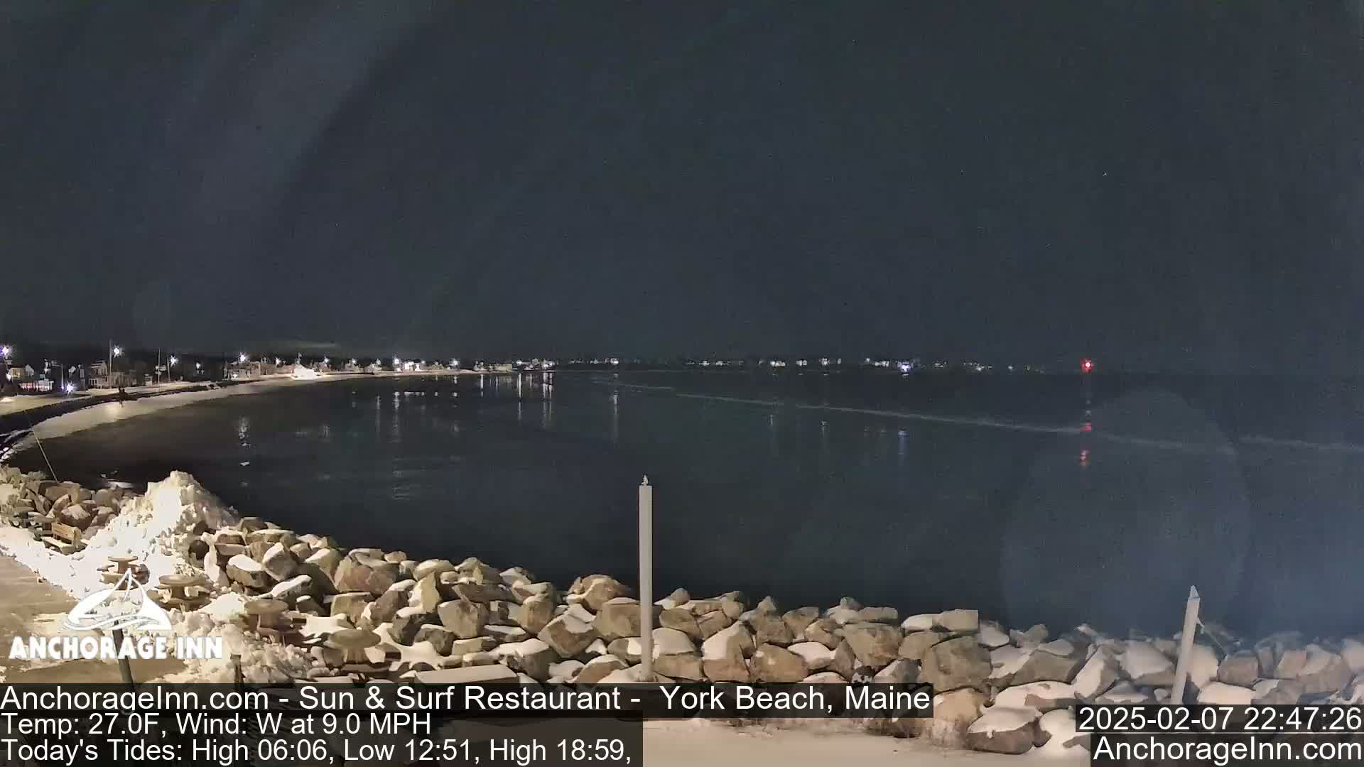 A nighttime view of a snow-dusted rocky shoreline and calm dark water, with a town faintly visible in the distance under a clear, dark sky.