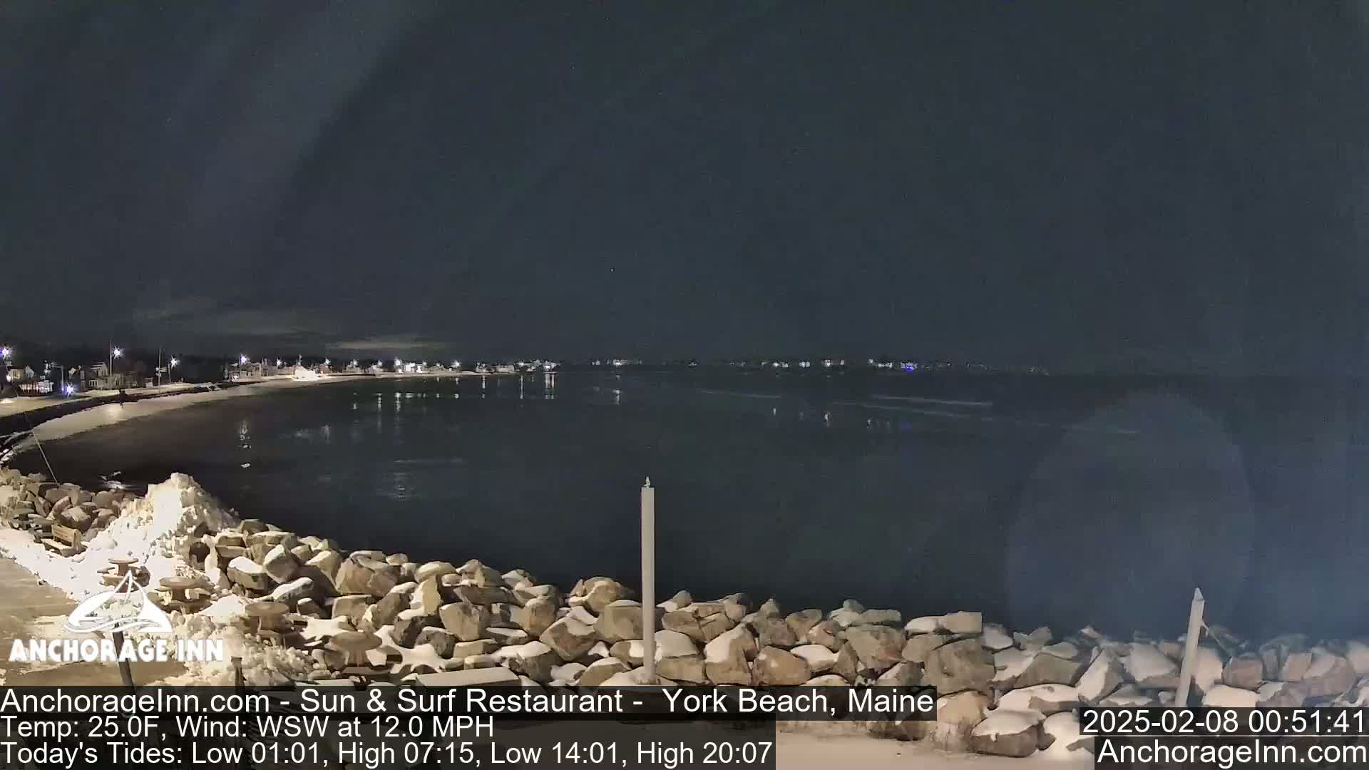 A nighttime view of a snow-dusted rocky shoreline and calm, dark water, with a town's lights visible in the distance under a clear, dark sky.