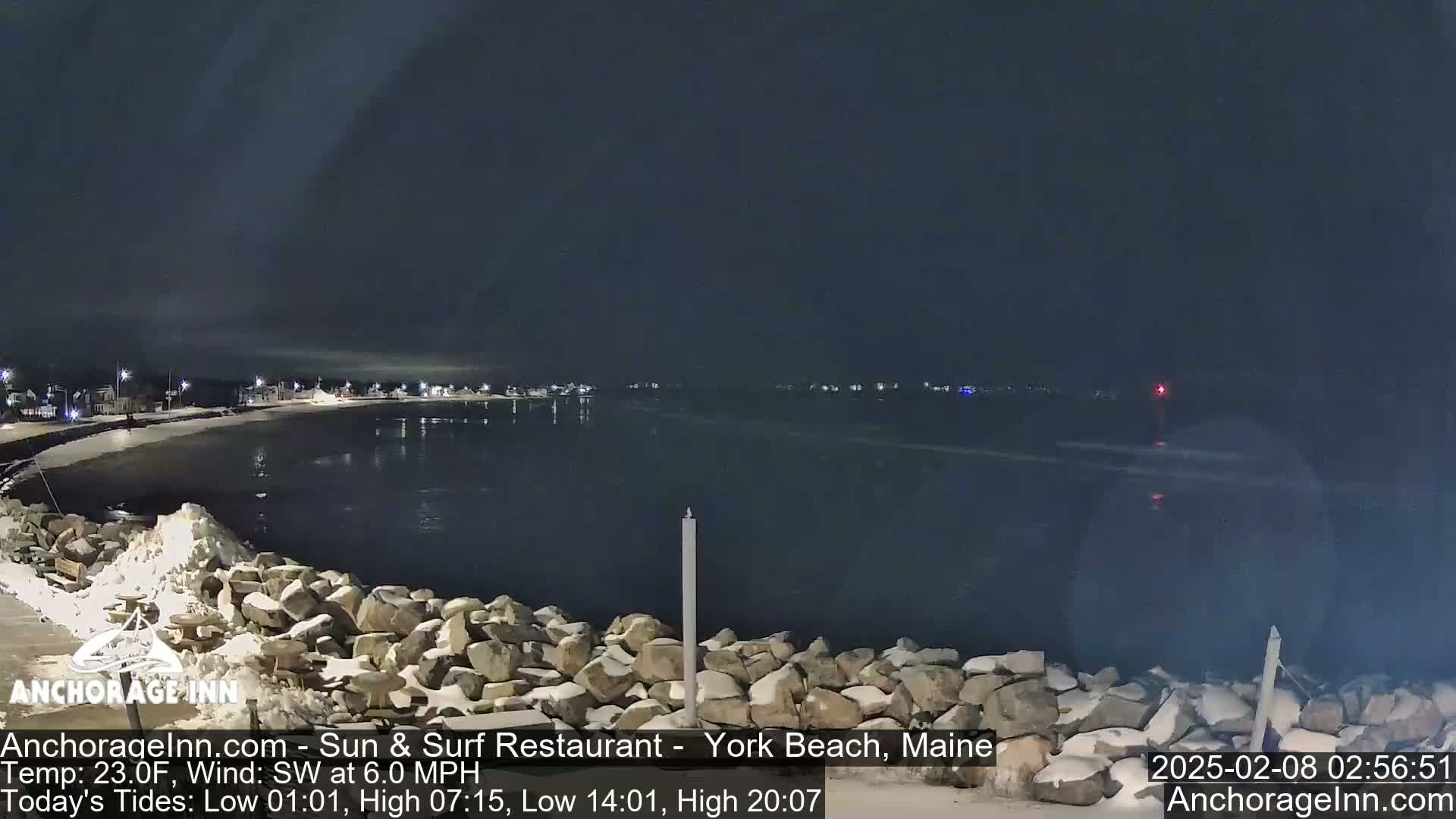 A snow-dusted rocky shoreline at night, overlooking a calm, dark body of water with faintly visible lights across the bay under a dark sky.