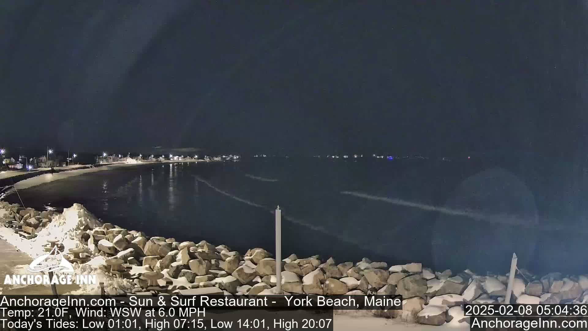 A nighttime view of a snow-covered rocky shoreline and calm dark water, with a few distant lights visible across the water under a clear dark sky.