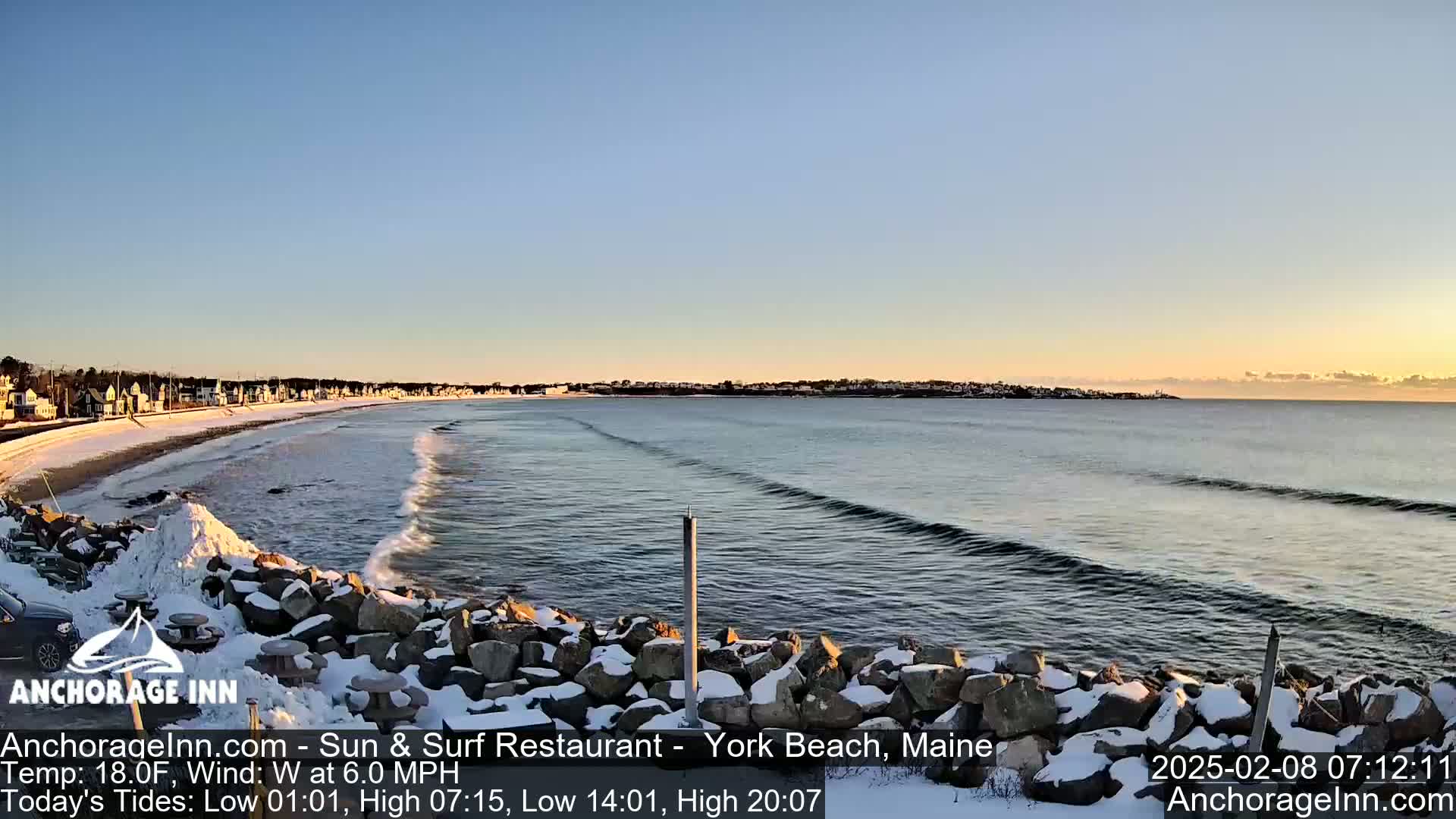 A snow-covered rocky shoreline overlooks a calm ocean with small waves under a clear, sunrise sky.