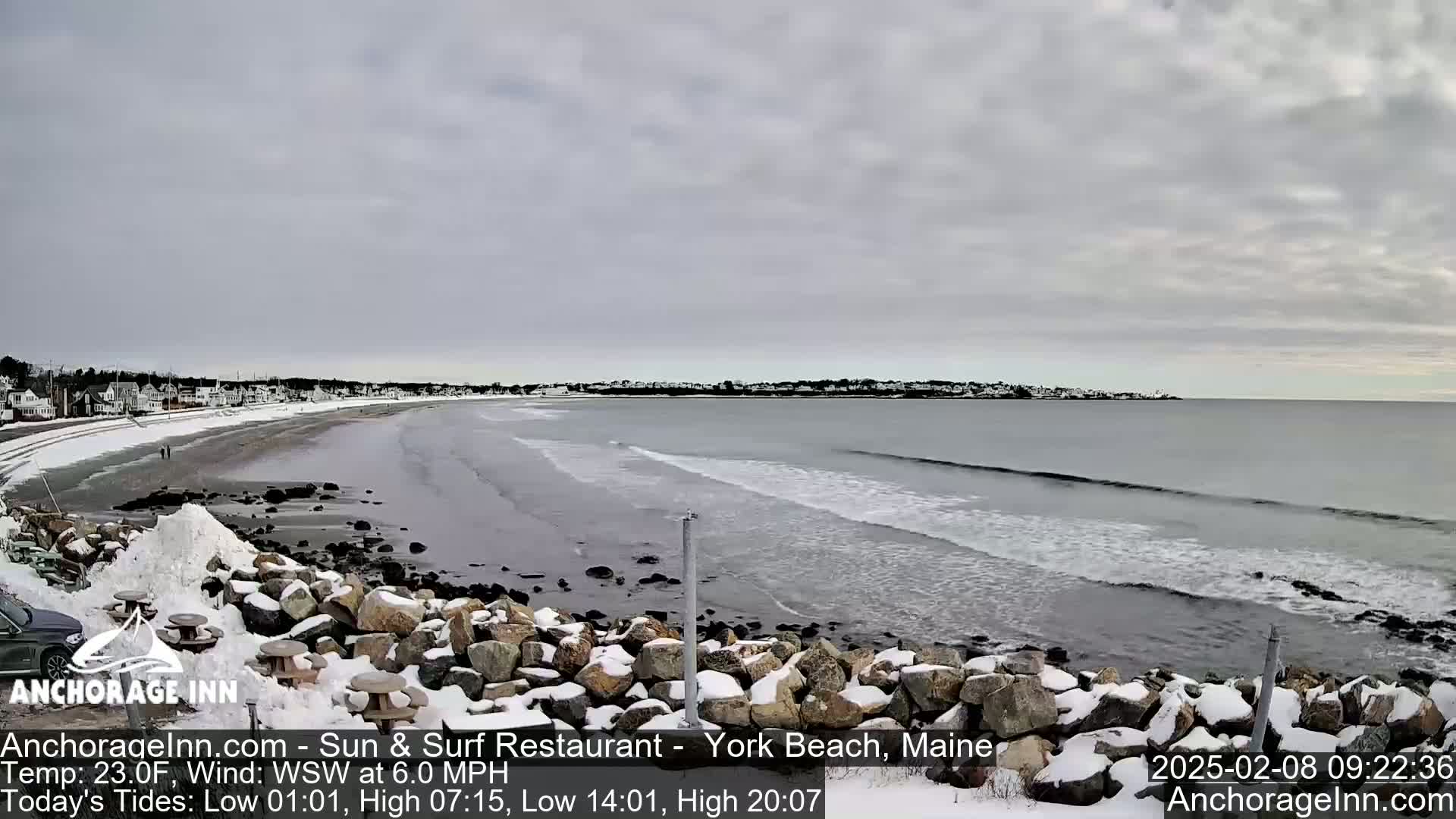 A snow-covered rocky shoreline with a partly cloudy sky overlooks a calm ocean with small waves.