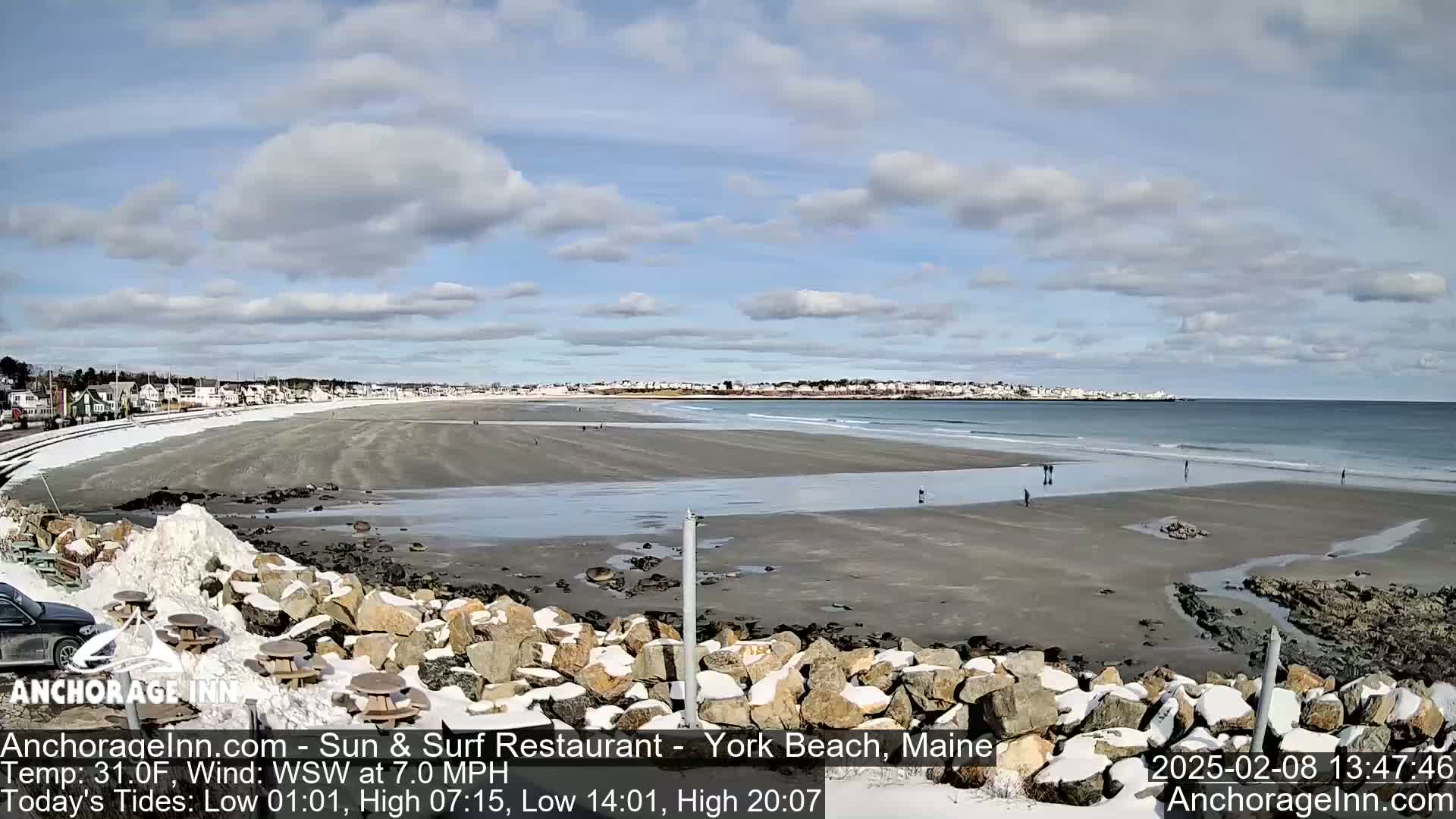 A sandy beach with a few people walking on it, partially covered in snow, stretches out to a calm ocean under a partly cloudy sky.