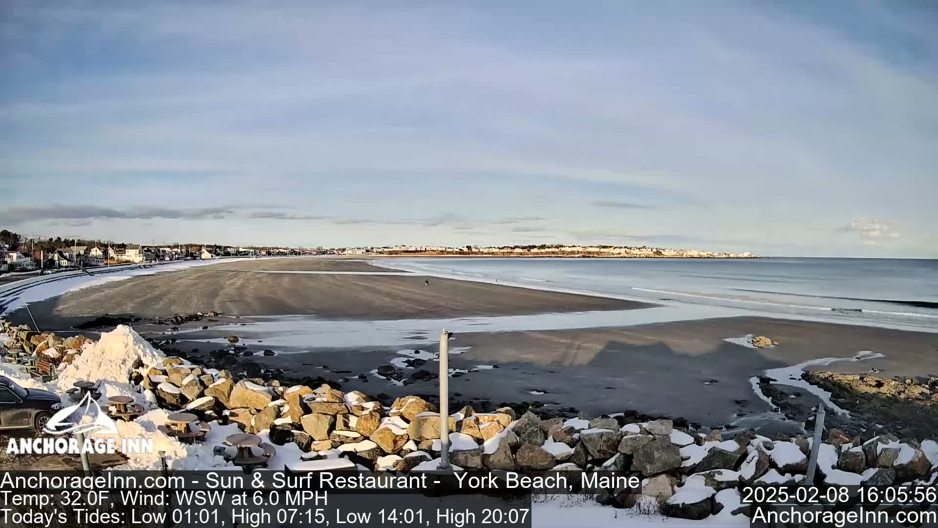 A partially snowy beach with calm waves stretches to a town under a clear, bright sky.