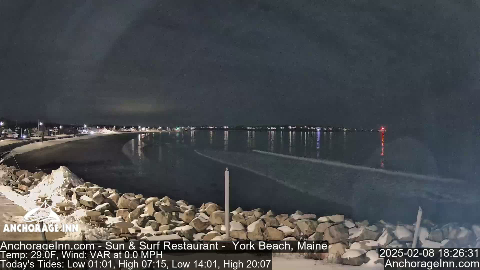 A nighttime view of a snow-dusted rocky shoreline and calm ocean, with a town faintly visible in the distance under a dark sky.