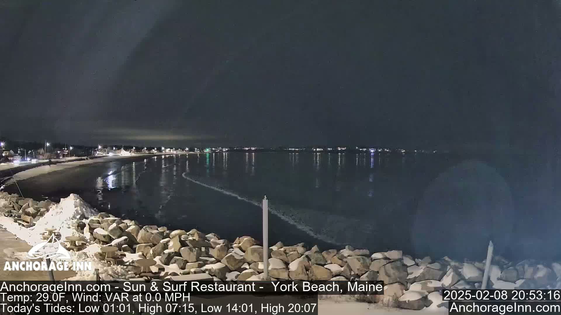 A nighttime view of a snow-dusted rocky shoreline and calm ocean reflecting the lights of a distant town under a dark sky.