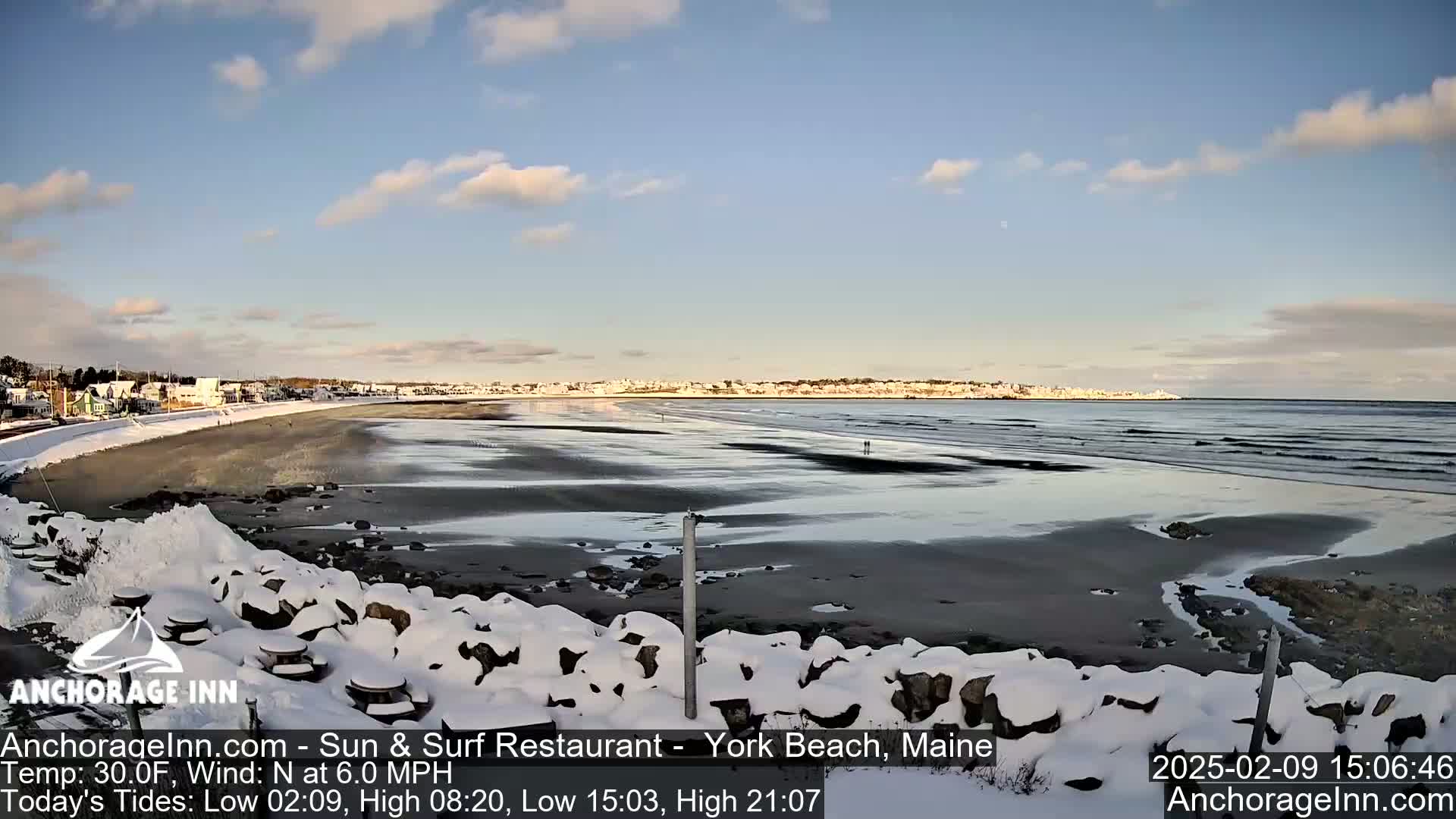 A snow-covered beach scene shows a partially frozen ocean with a town in the distance under a partly cloudy sky.