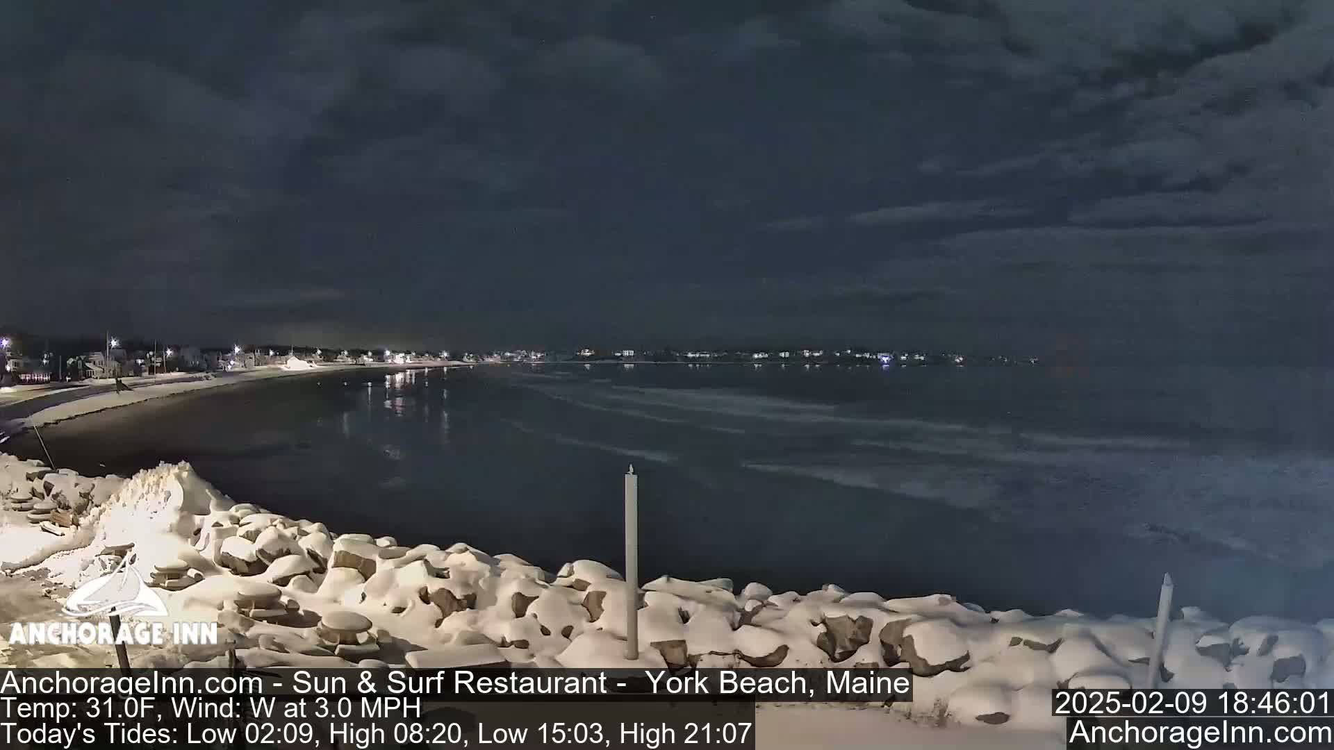 A snow-covered rocky shoreline at night overlooks a dark, wavy ocean under a cloudy sky.