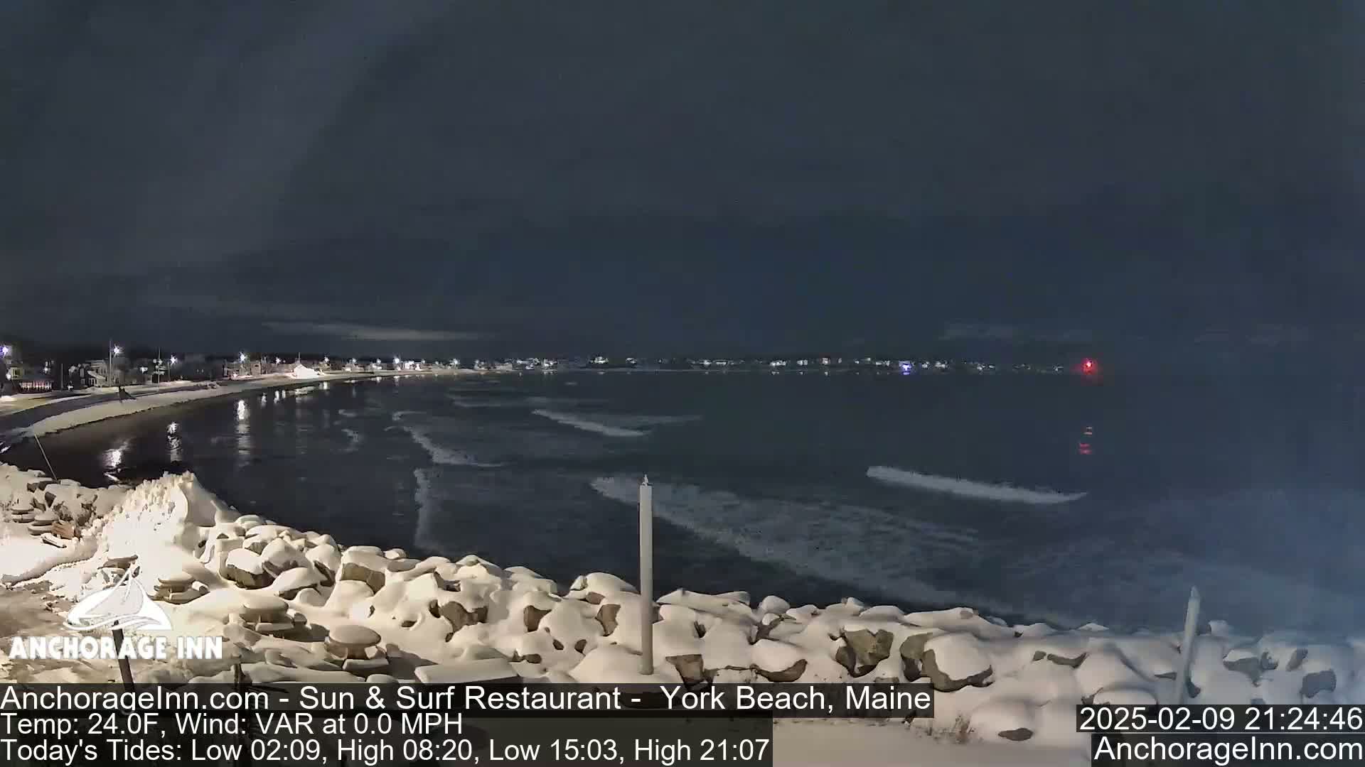 A snow-covered rocky shoreline at night overlooks a dark ocean with small waves under a clear, dark sky.