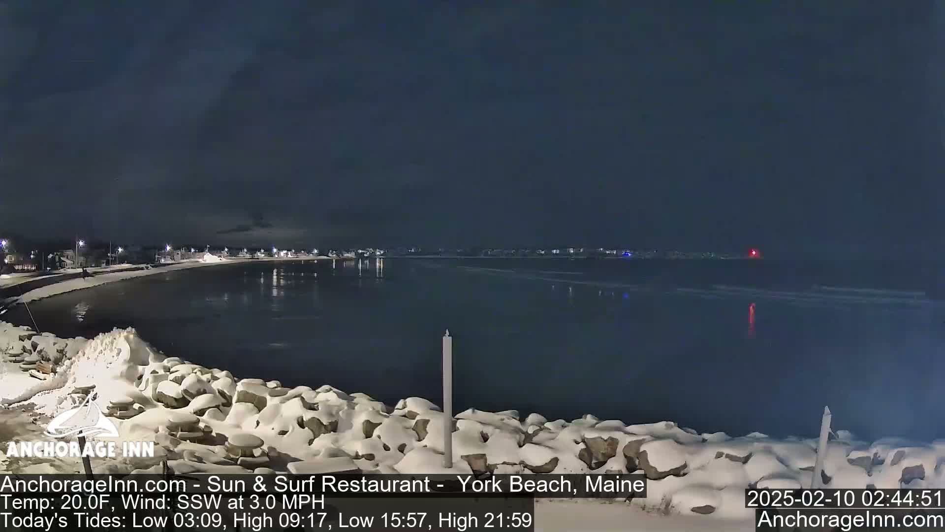 A snow-covered rocky shoreline at night overlooks a dark, calm body of water with a town faintly visible in the distance under a clear, dark sky.