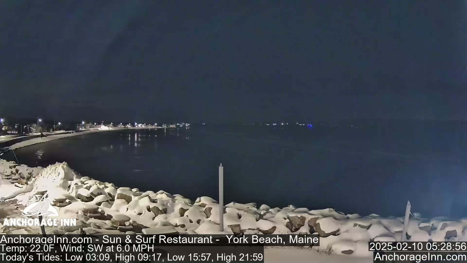 A snow-covered rocky shoreline at night overlooks a calm, dark body of water under a clear, dark sky.