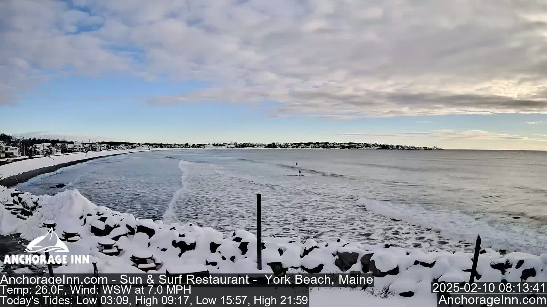 A snow-covered rocky shoreline overlooks a gray ocean with small waves under a partly cloudy sky.