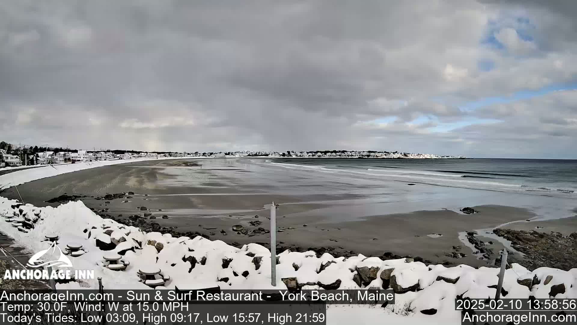A snow-covered rocky shoreline overlooks a beach with gentle waves under a cloudy sky.
