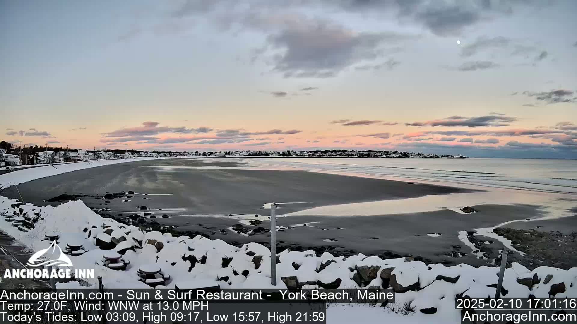 A snow-covered rocky shoreline overlooks a calm ocean beach at sunset under a partly cloudy sky.