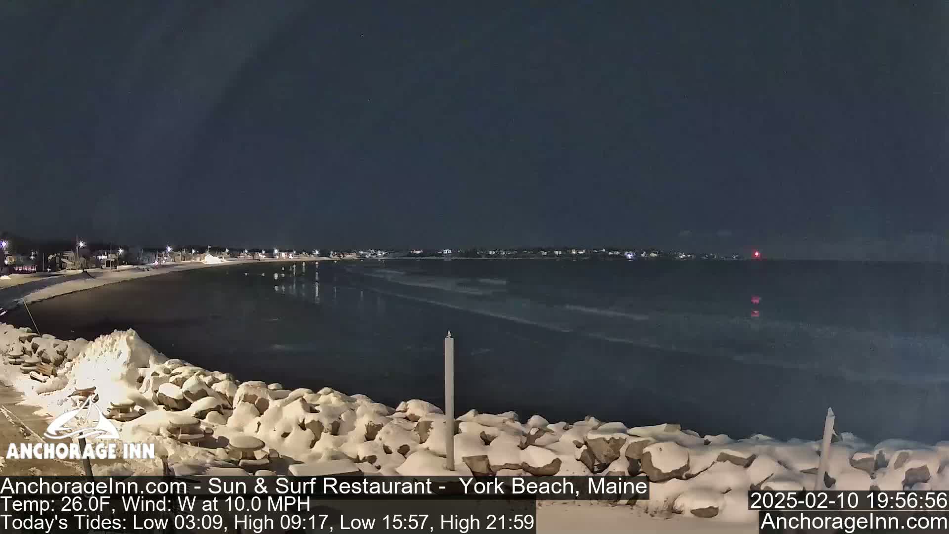 A snow-covered rocky shoreline at night overlooks a dark, wavy ocean under a clear sky.