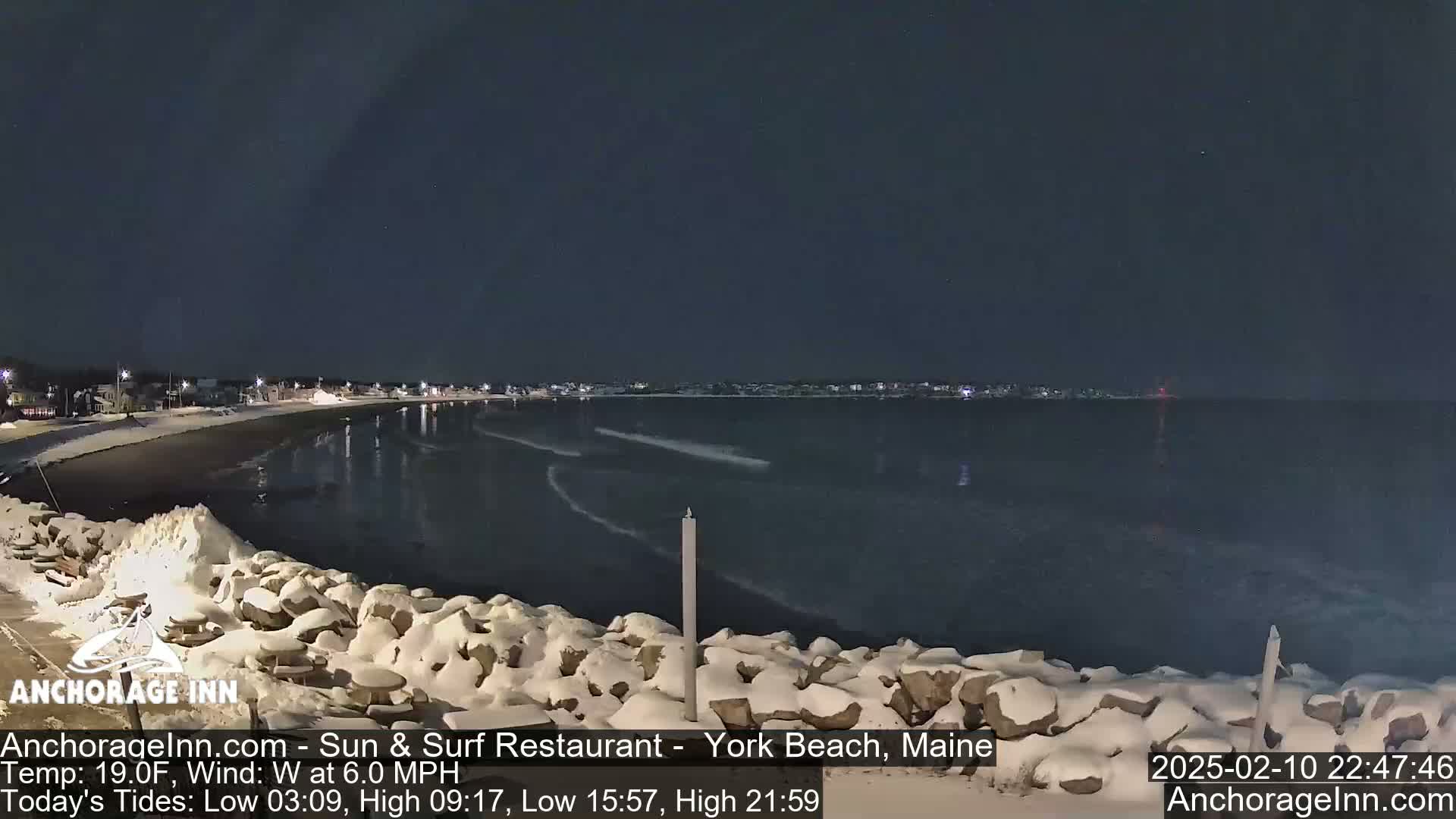 A snow-covered rocky shoreline at night overlooks a dark, calm ocean with a town visible in the distance under a clear, dark sky.