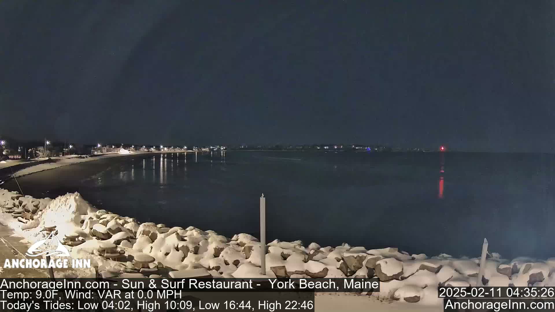 A snow-covered rocky shoreline at night overlooks a calm, dark body of water under a clear, dark sky.