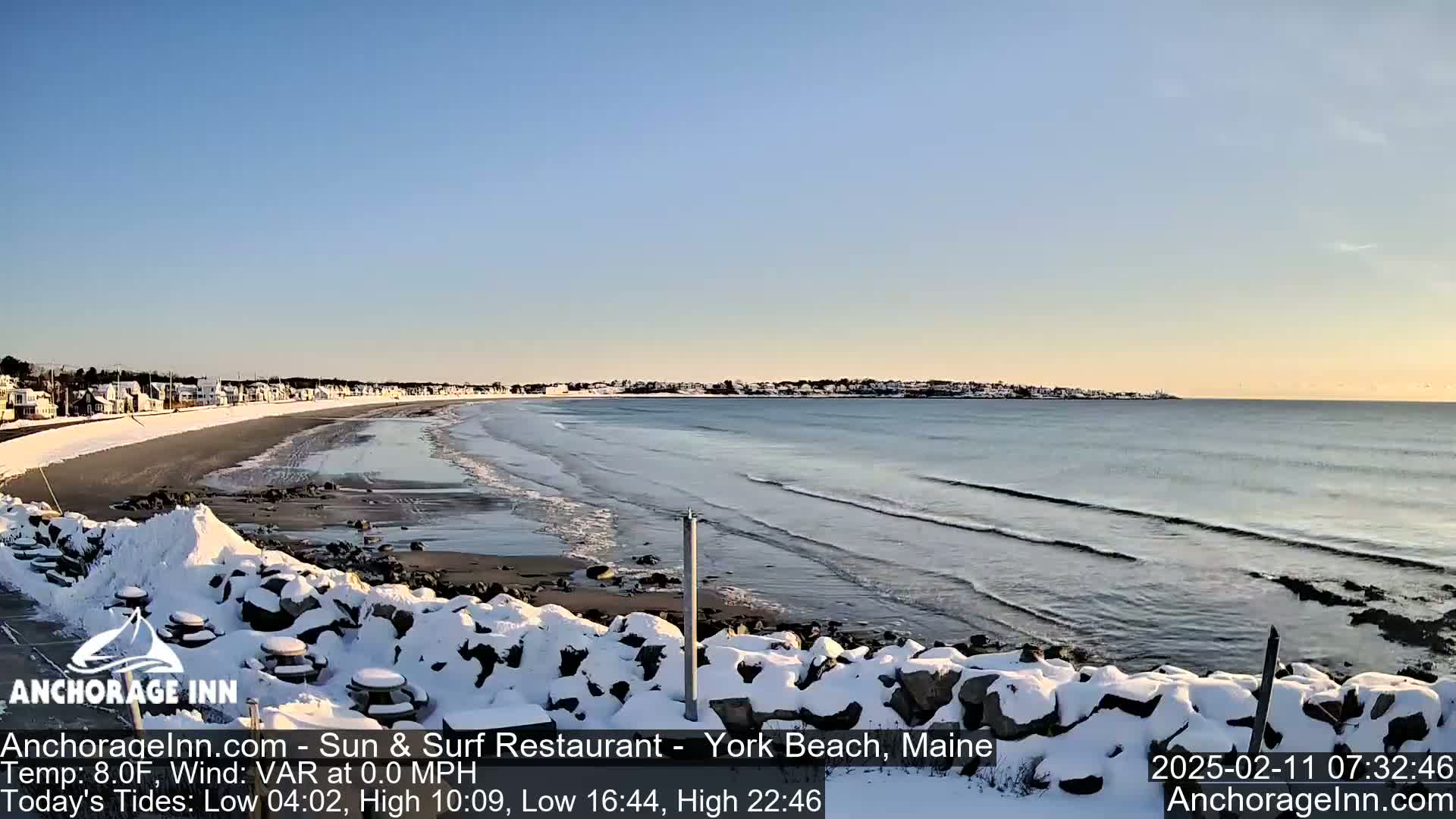 A snow-covered rocky shoreline overlooks a calm ocean with gentle waves under a clear, light blue sky.