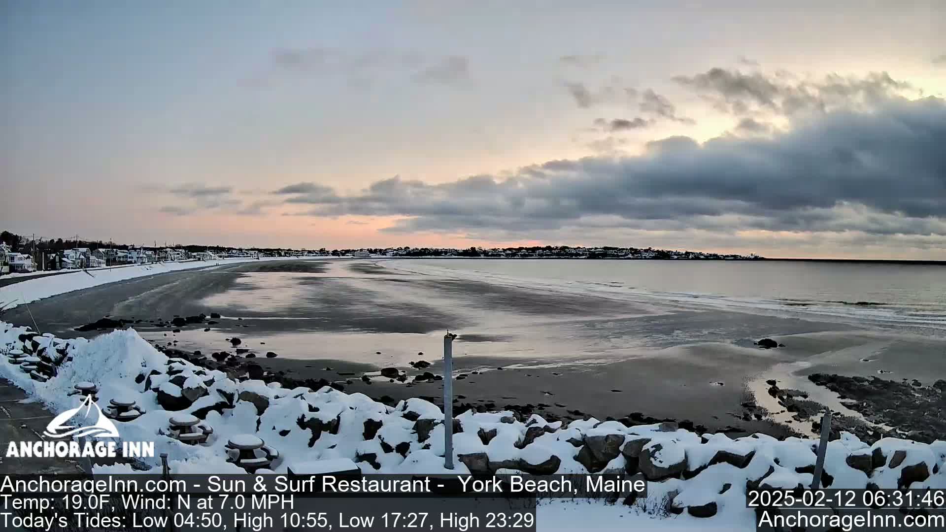 A snow-covered rocky shoreline overlooks a calm, dark gray ocean under a partly cloudy sky at sunrise.