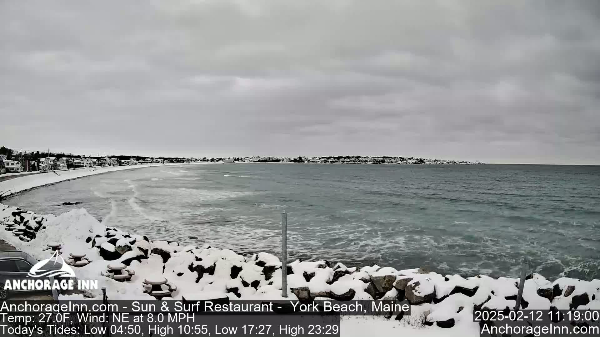 A snow-covered rocky shoreline and beach overlook a gray, wavy ocean under a cloudy sky.