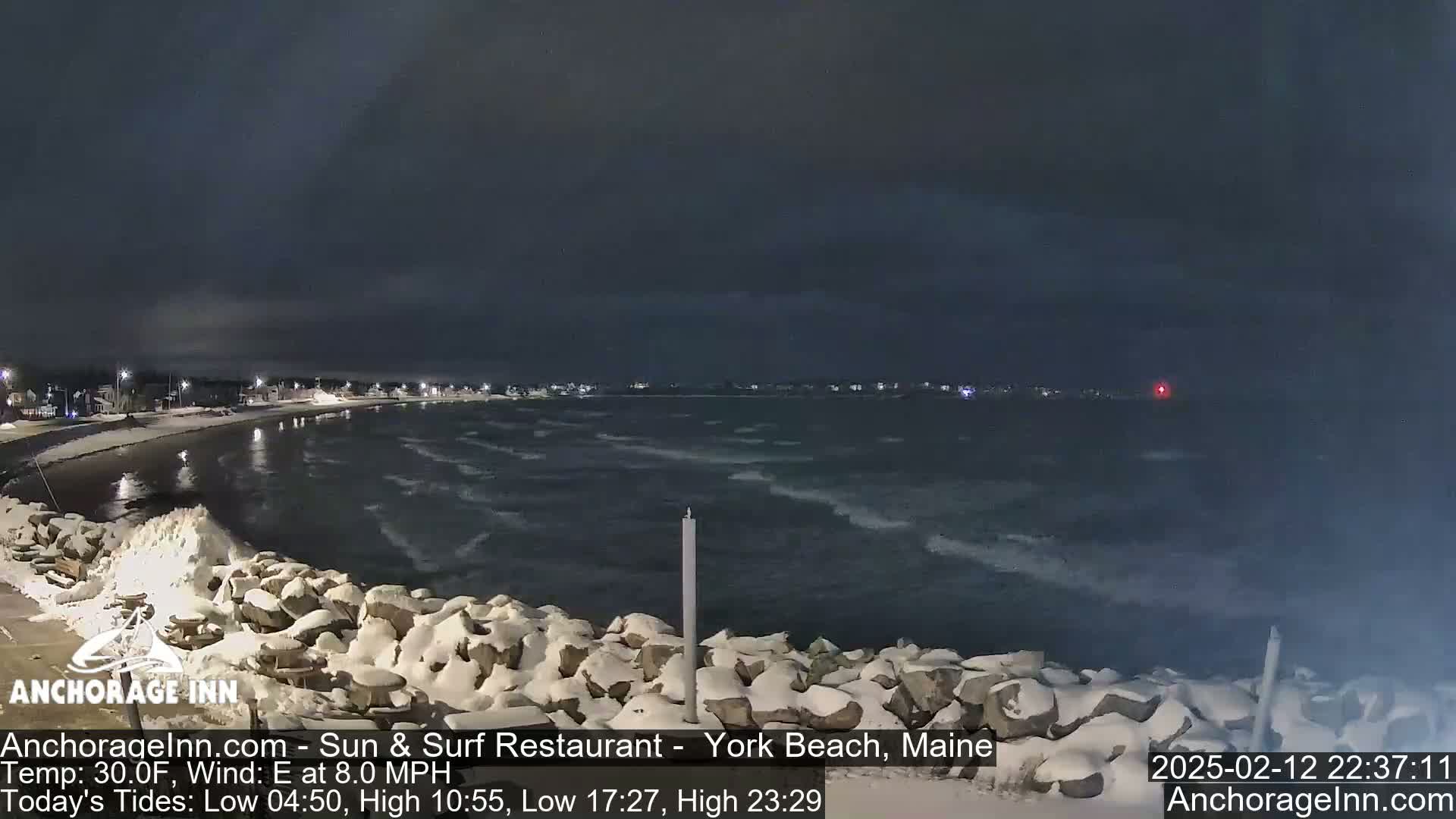 A snow-covered rocky shoreline at night overlooks a dark, choppy ocean under a cloudy sky.