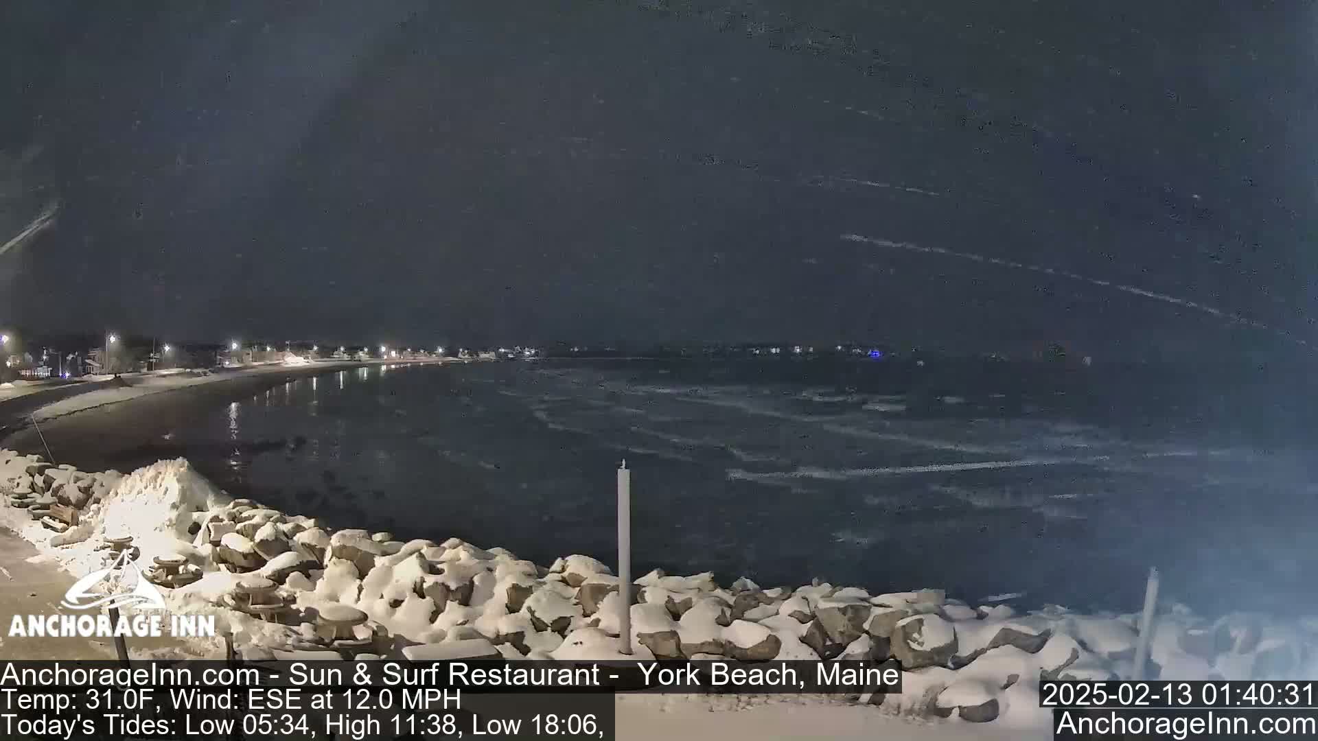 A snow-covered rocky shoreline at night with a dark, choppy sea under a lightly snowing sky.