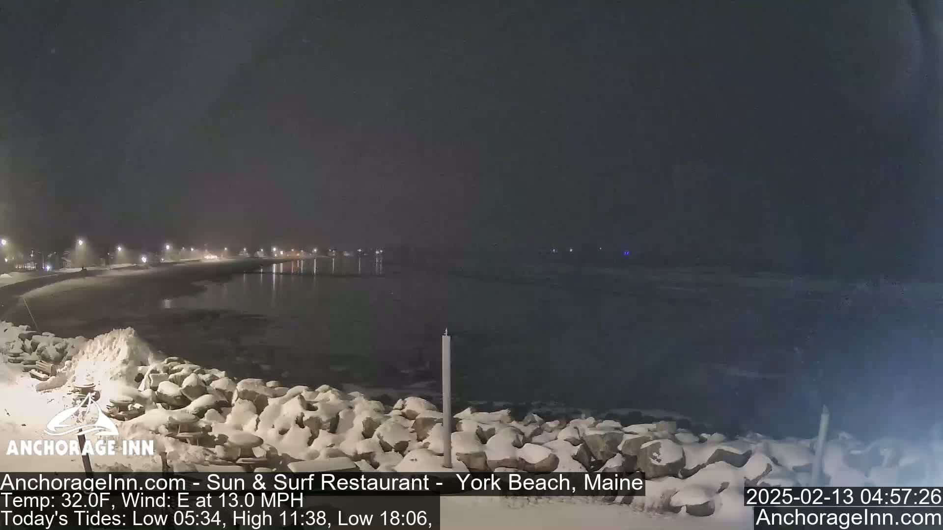 A snow-covered rocky shoreline at night, with a calm, dark body of water and faintly lit buildings in the distance under a dark, clear sky.