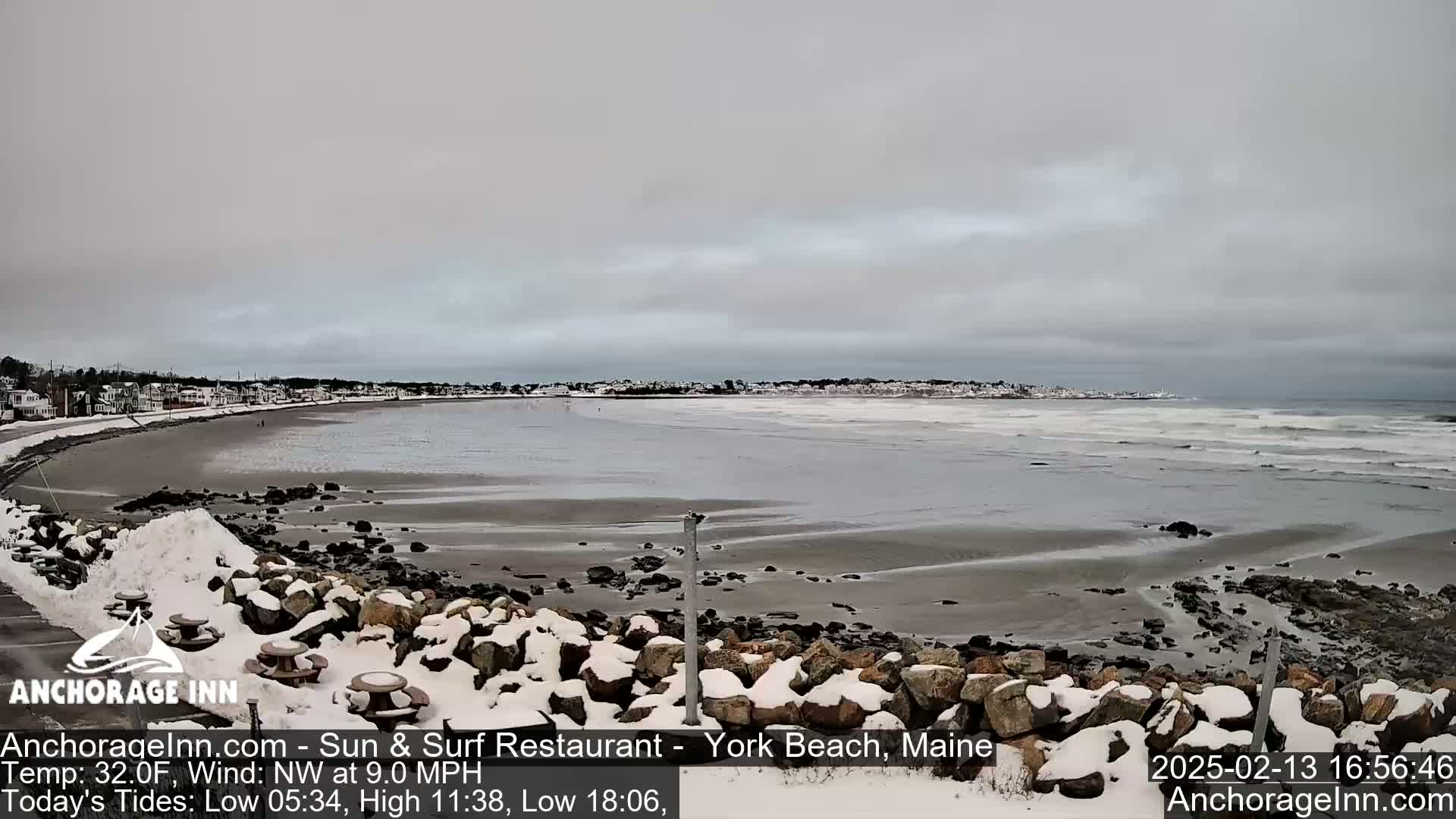 A snowy shoreline with a rocky embankment overlooks a gray, overcast ocean with small waves.
