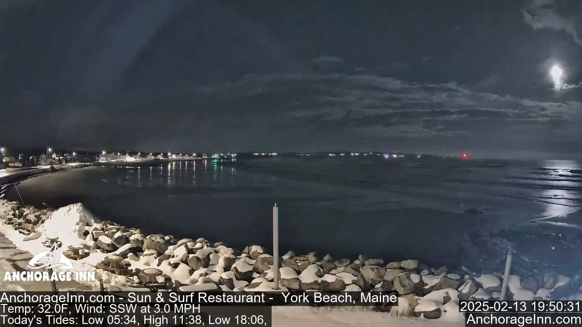 A moonlit nighttime scene shows a snow-covered rocky shoreline next to a calm, dark body of water with a town faintly visible in the distance under a partly cloudy sky.