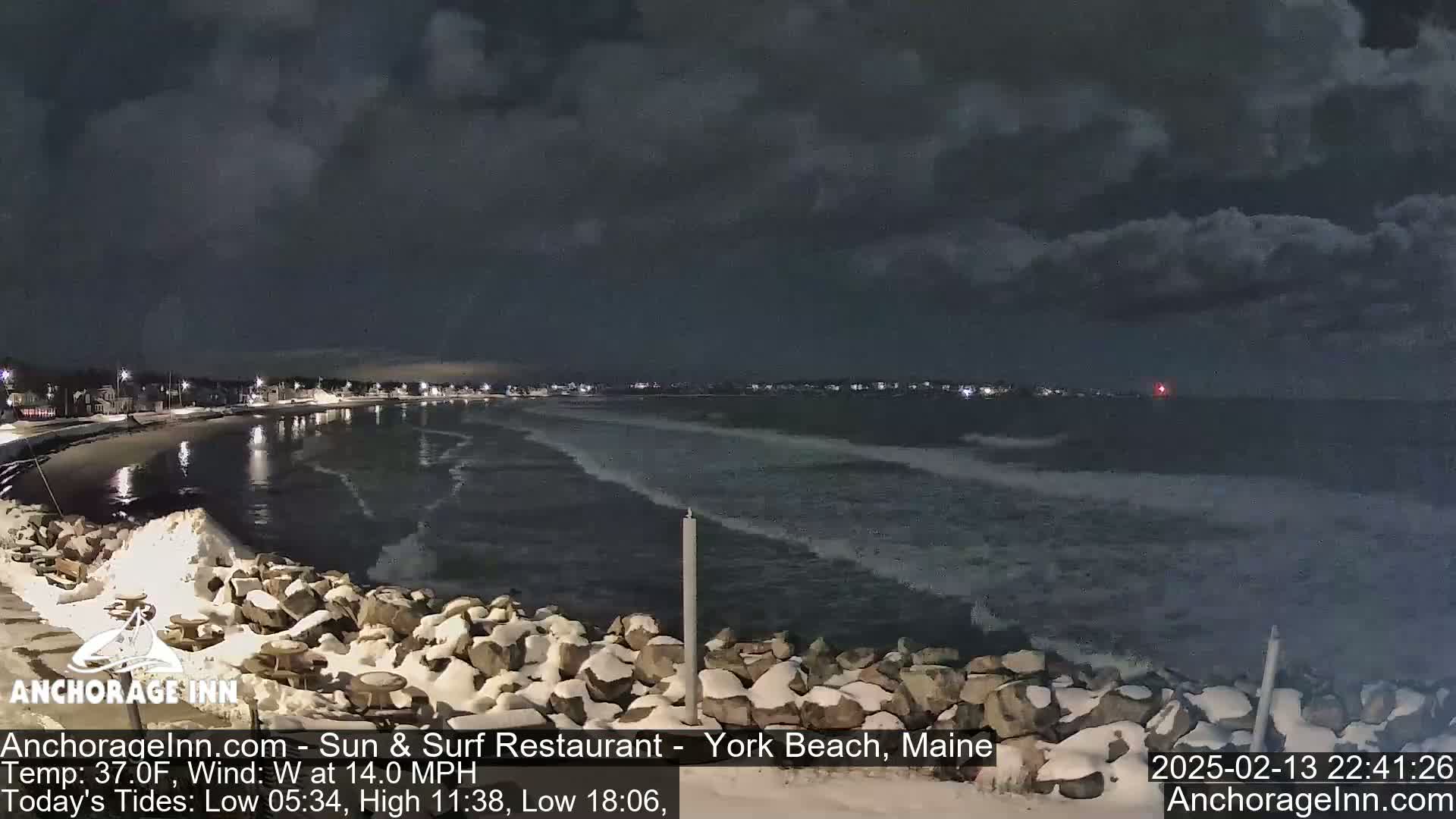 A snow-dusted rocky shoreline at night shows a dark, wavy ocean under a cloudy sky.