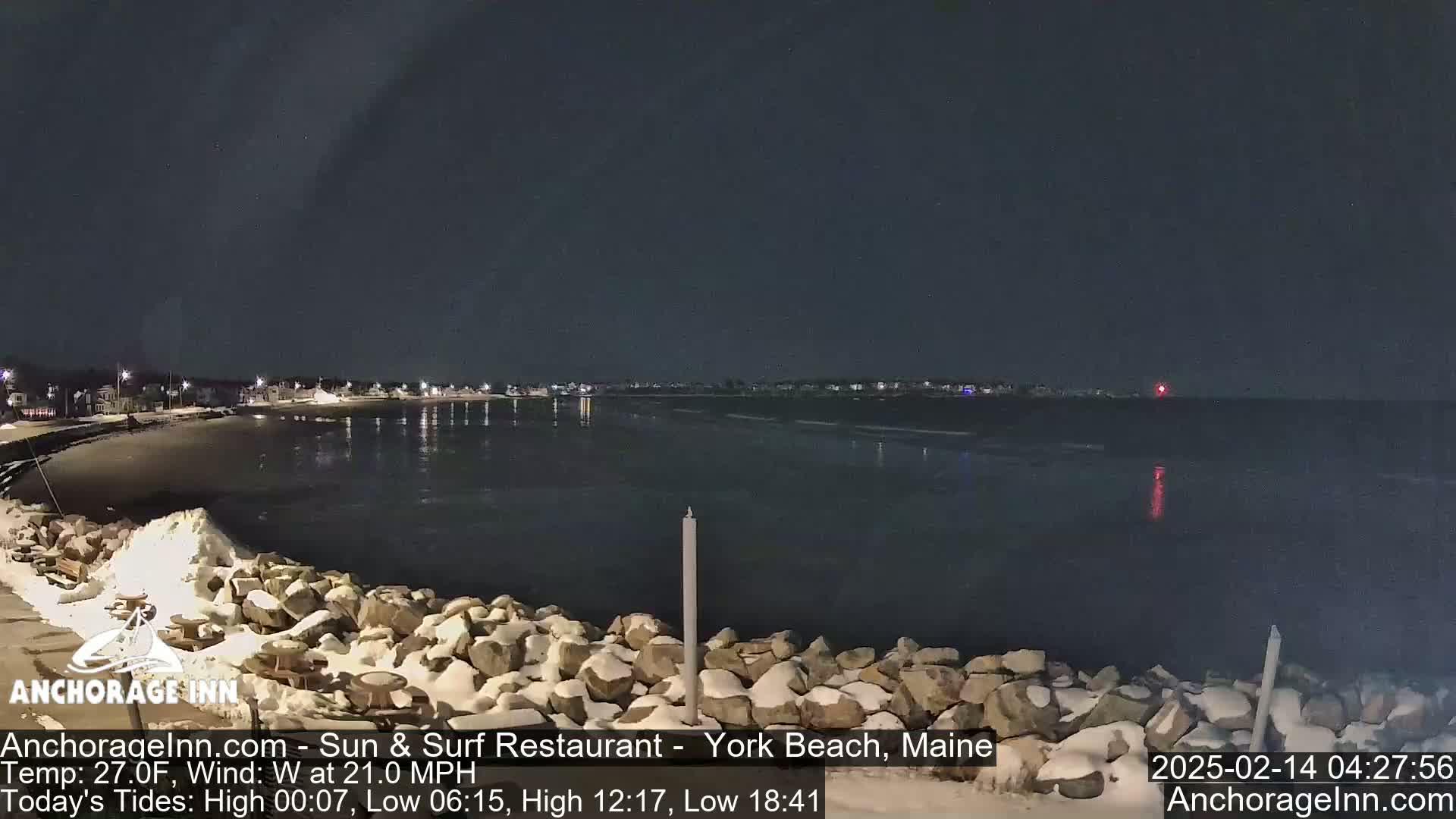 A nighttime view of a snow-dusted rocky shoreline and calm dark water, with a town faintly lit in the distance under a clear, dark sky.
