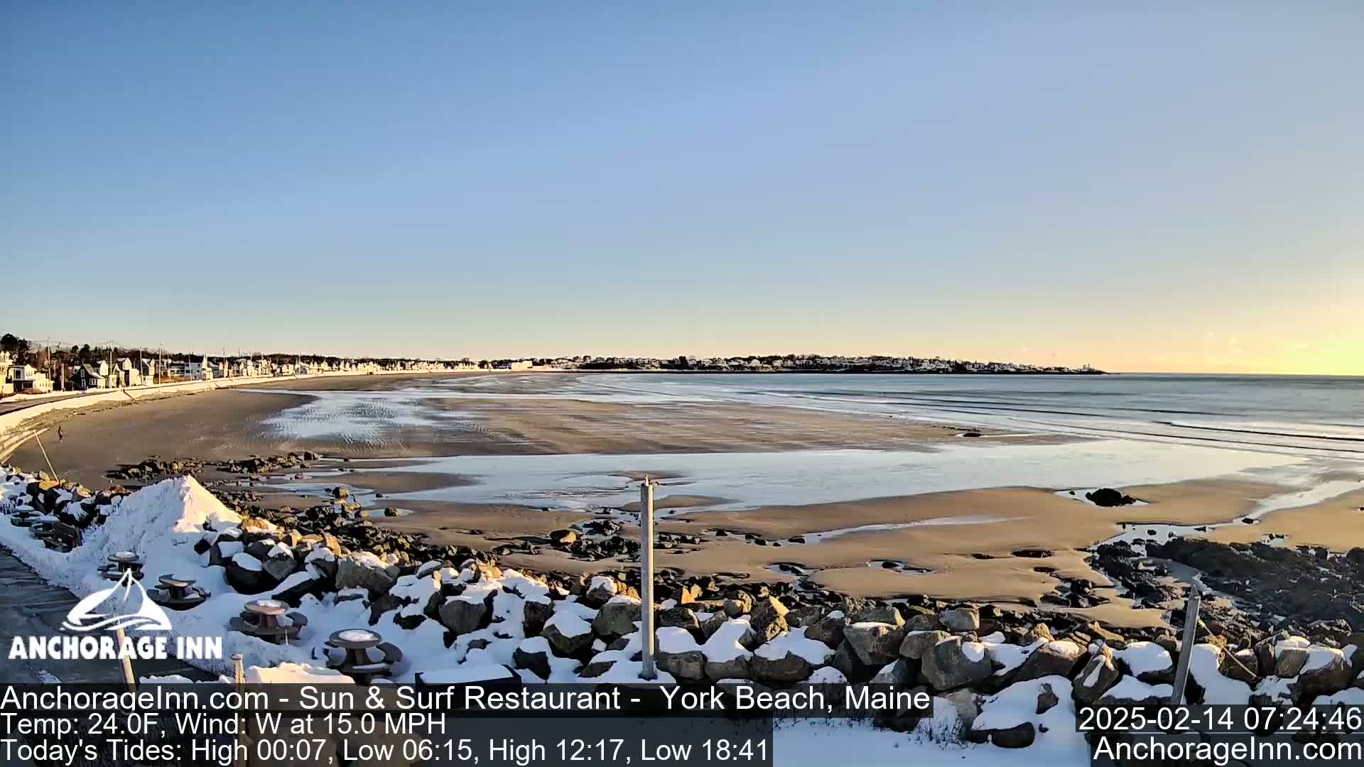 A snow-dusted beach with calm waves and a clear blue sky stretches out to a distant town under a partly sunny sky.