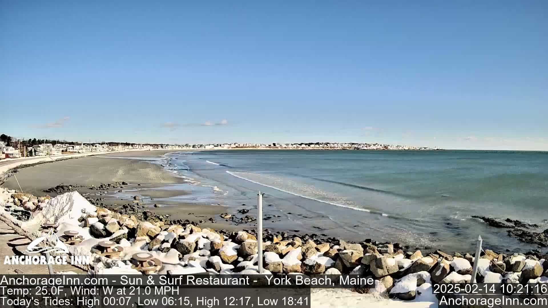 A partially snow-covered rocky shoreline overlooks a calm ocean with small waves under a clear blue sky.