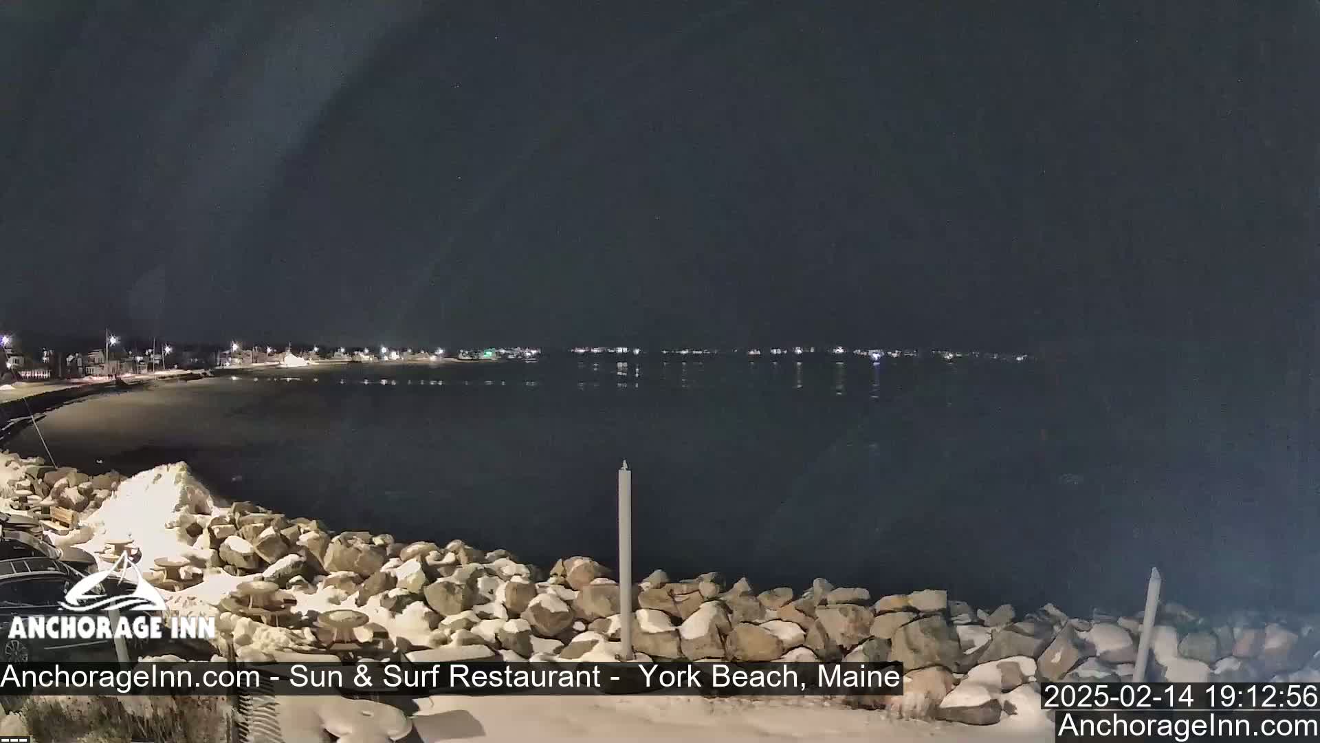 A nighttime view of a snow-dusted rocky shoreline and calm water, with a faintly lit town visible in the distance under a dark sky.