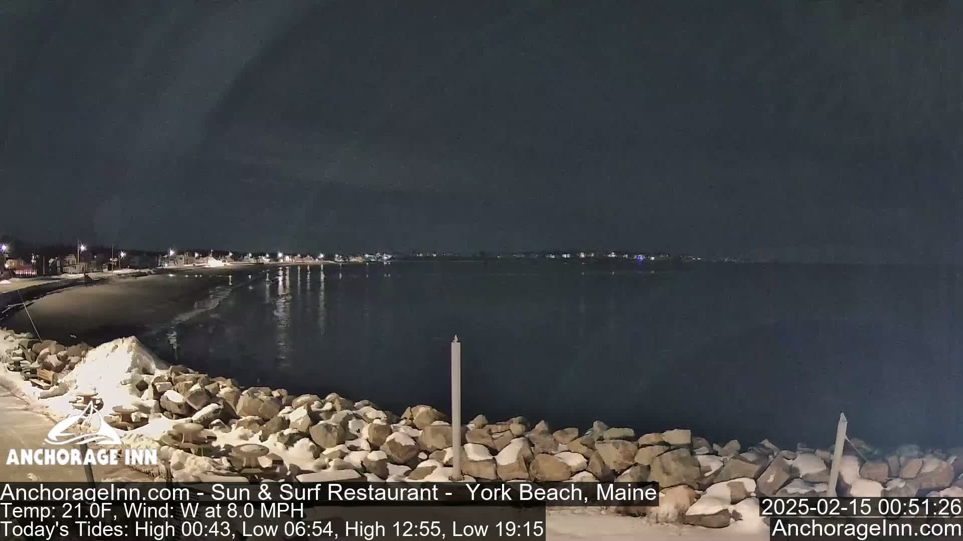 A nighttime view of a snow-dusted rocky shoreline and calm ocean under a dark sky with a slight wind.