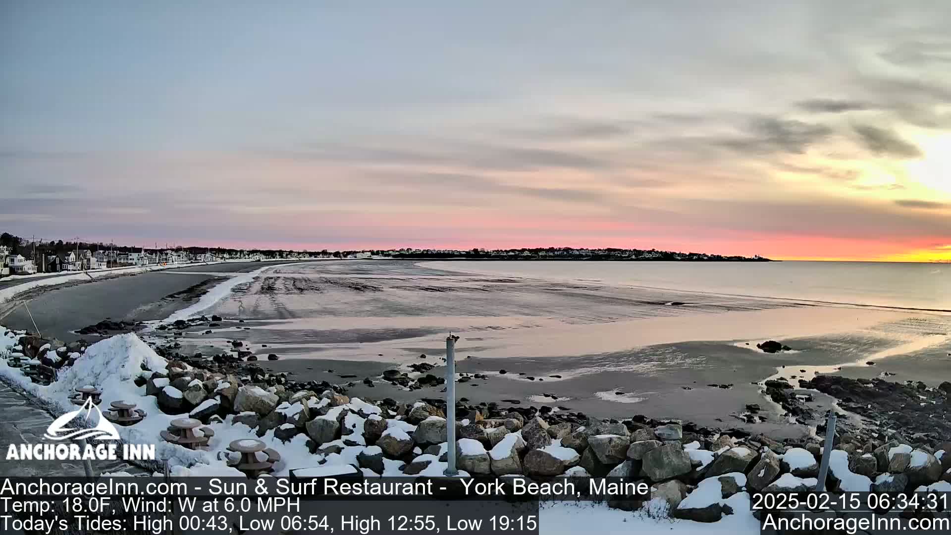 A partly cloudy sunrise over a snow-dusted rocky beach with a calm ocean and a town visible in the distance.