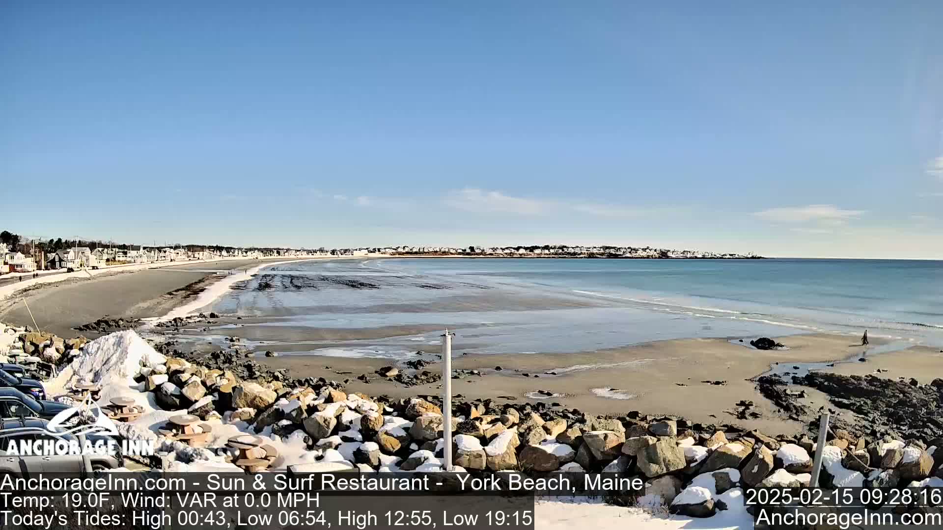 A mostly sunny day shows a sandy beach with some snow-covered rocks in the foreground, and a calm ocean meeting a shoreline with buildings in the distance.