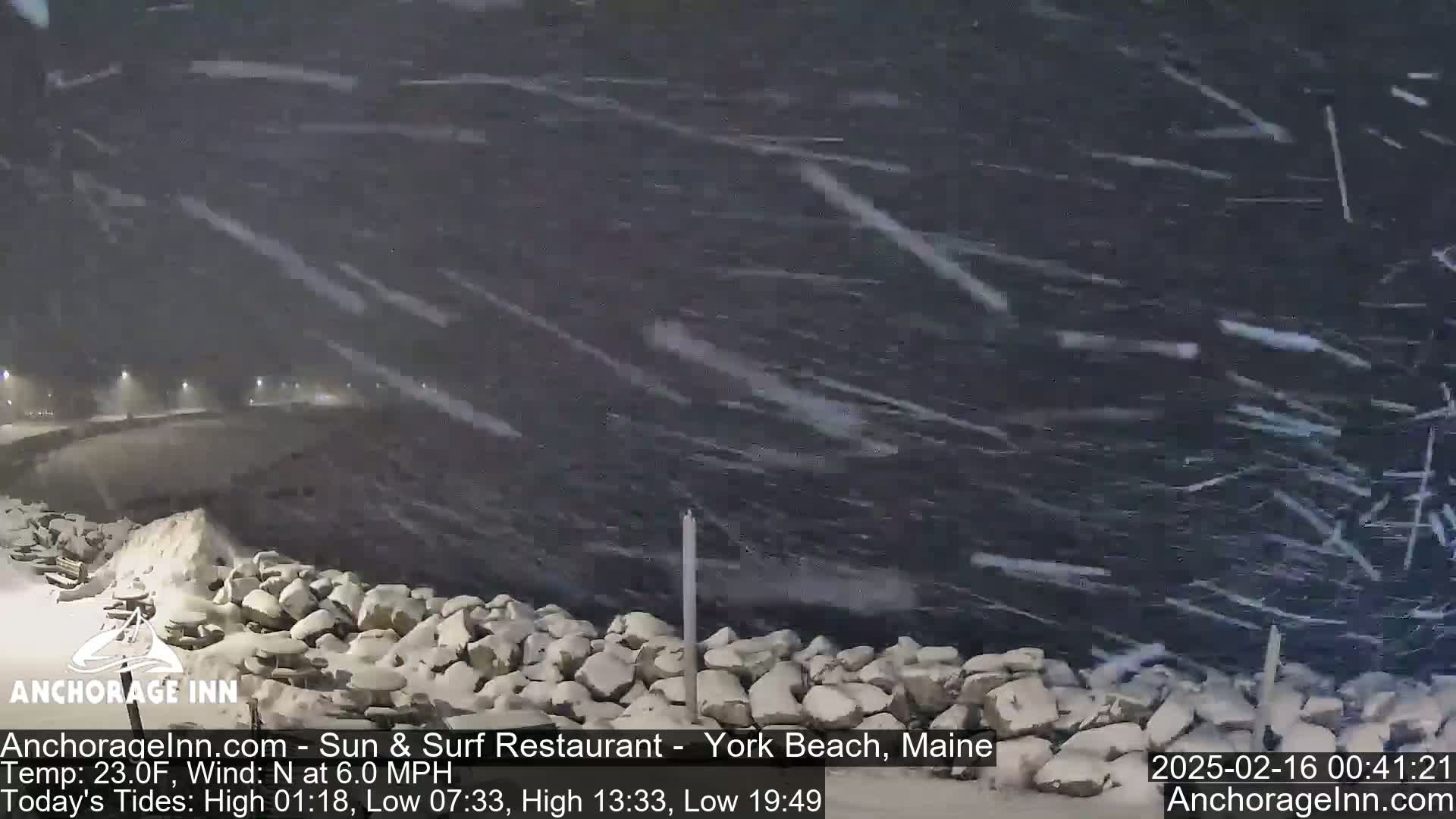 A snowstorm over a dark body of water, with snow-covered rocks and a roadway visible in the distance.