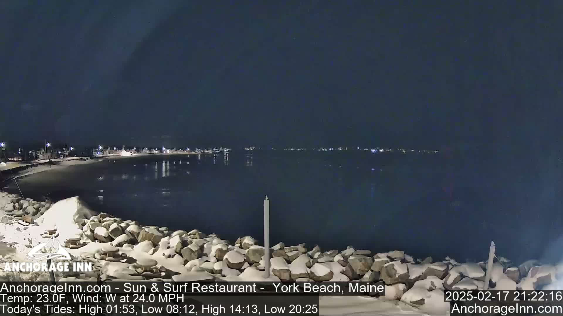 A snow-covered rocky shoreline at night overlooks a dark, calm body of water with a distant town illuminated by lights under a clear, dark sky.