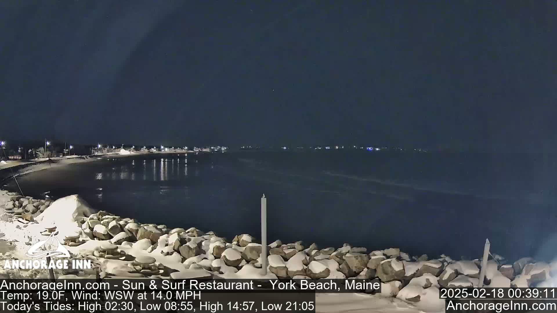 A snow-covered rocky shoreline at night overlooks a dark, calm body of water with a town faintly lit in the distance under a clear, dark sky.
