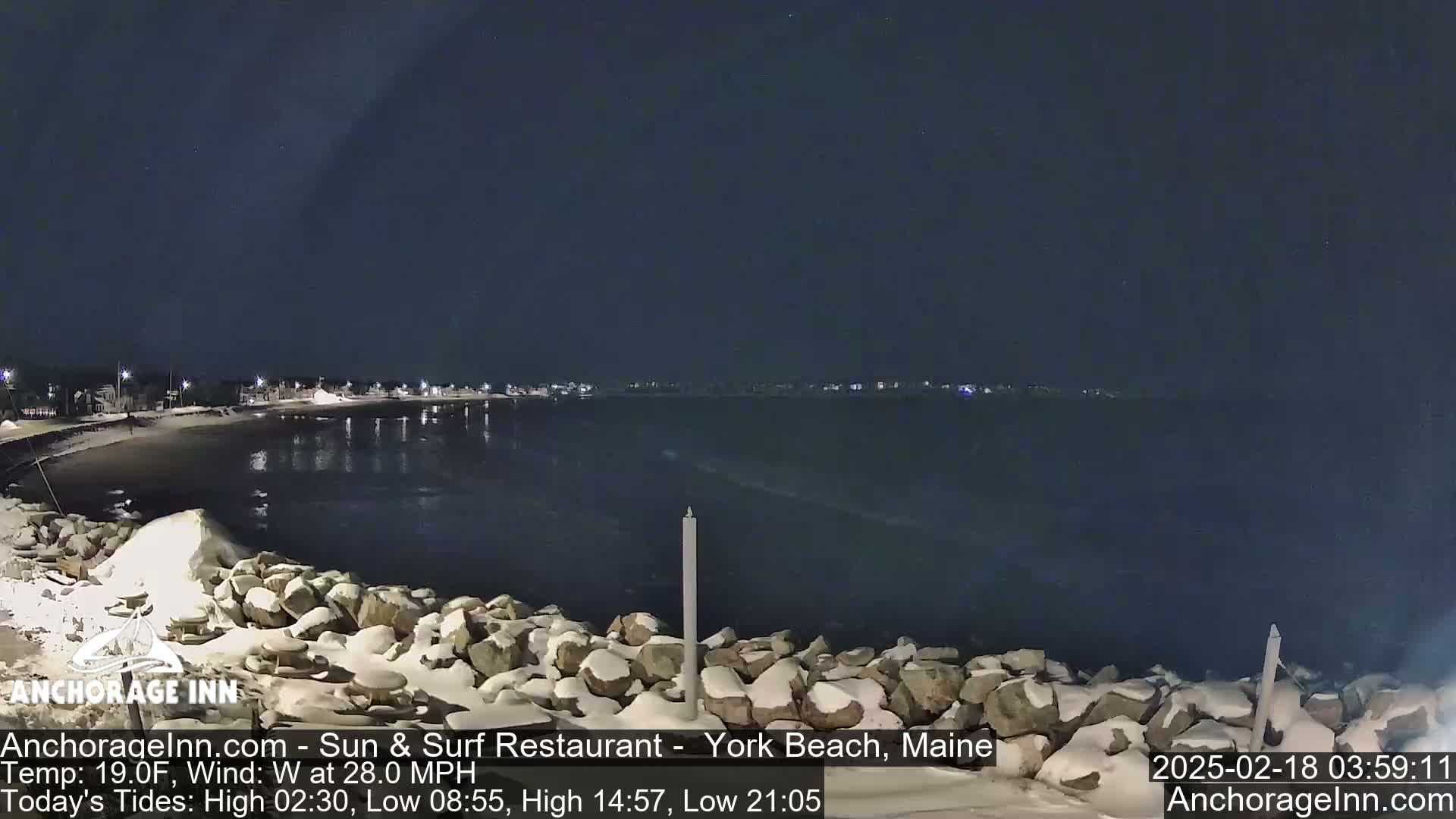 A snow-covered rocky shoreline at night overlooks a dark, wavy ocean under a clear sky with a 28 mph west wind.