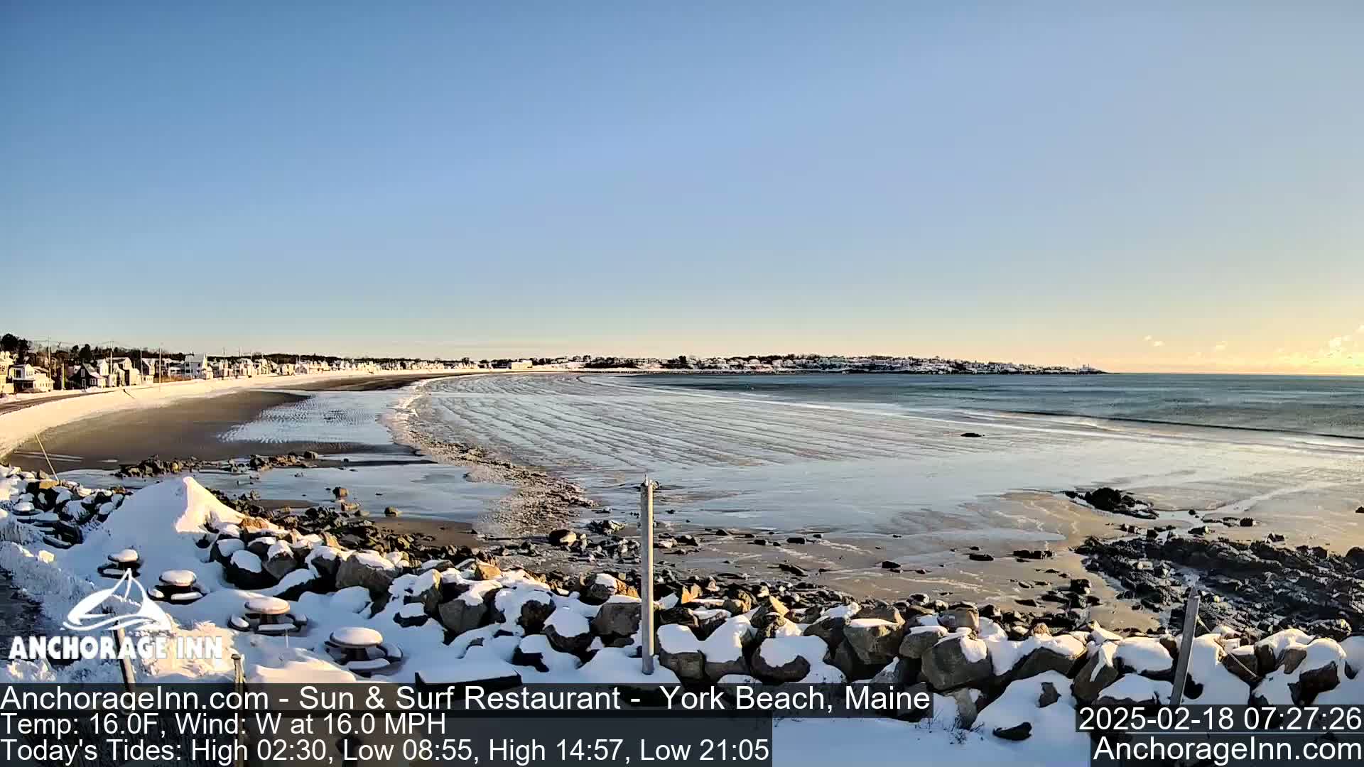 A snow-covered rocky shoreline overlooks a partly frozen sandy beach and calm ocean under a clear sky.