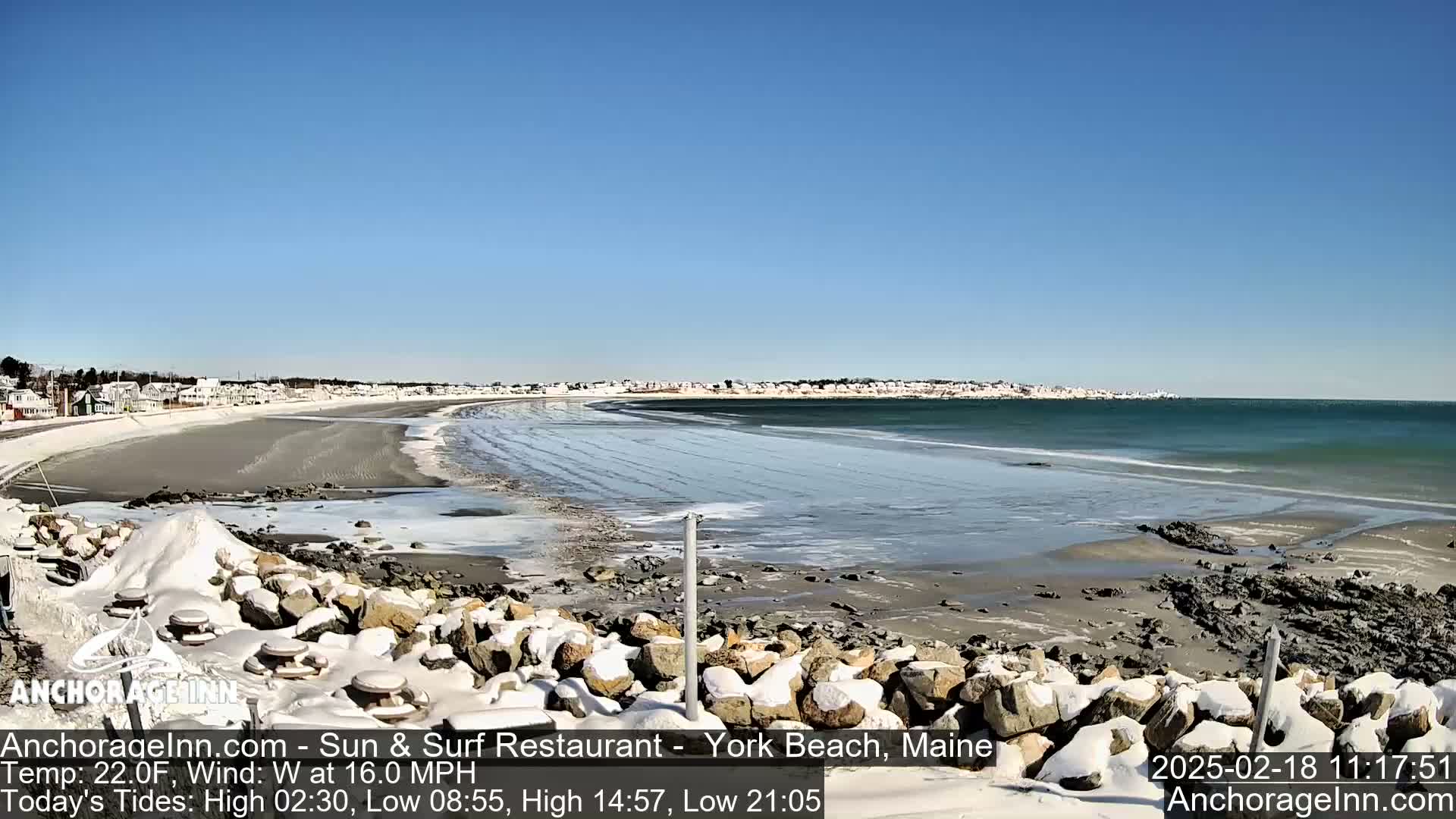 A snow-covered rocky shoreline borders a partially frozen sandy beach and ocean with a town in the background on a sunny, windy day.