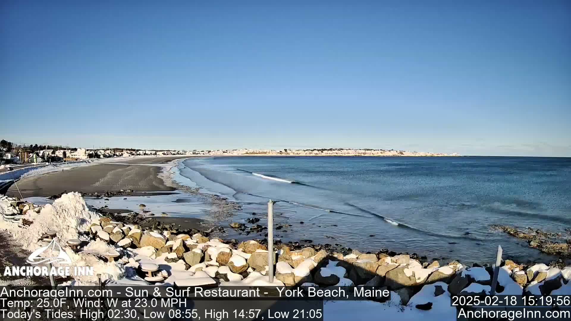 A snow-dusted beach with small waves and a town in the distance under a clear blue sky.