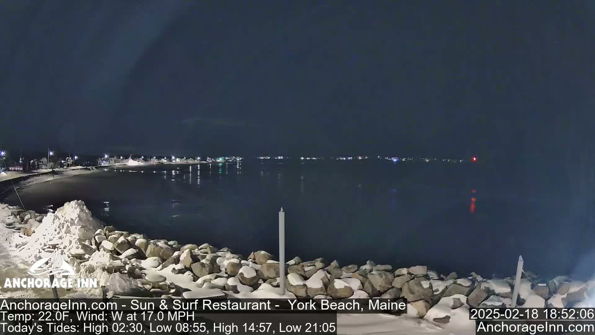 A snow-covered rocky shoreline at night overlooks a calm, dark body of water reflecting a distant town's lights under a clear sky.