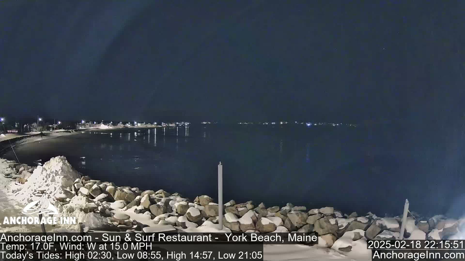 A snow-covered rocky shoreline at night overlooks a dark, calm body of water with a town faintly lit in the distance under a clear sky.