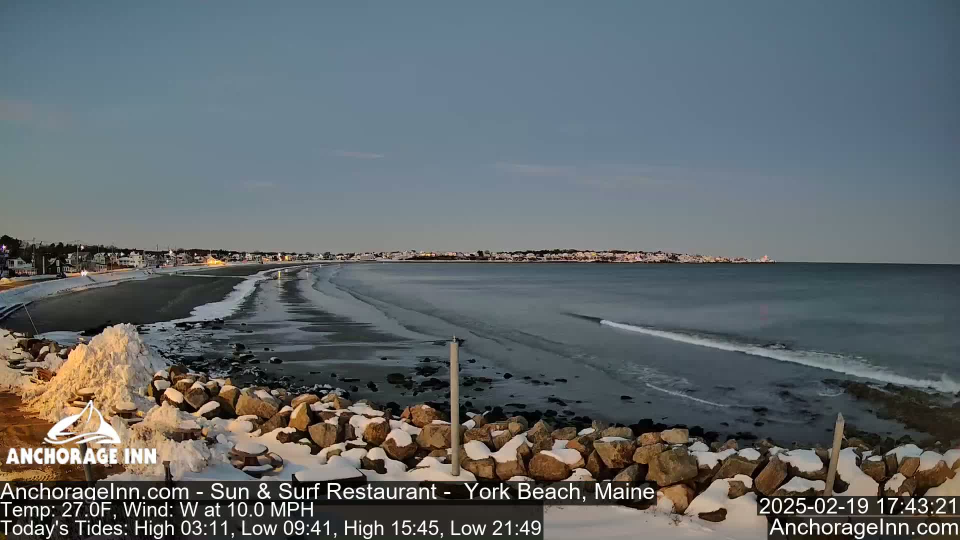 A snow-dusted rocky shoreline overlooks a calm, dark-blue ocean with gentle waves under a twilight sky.